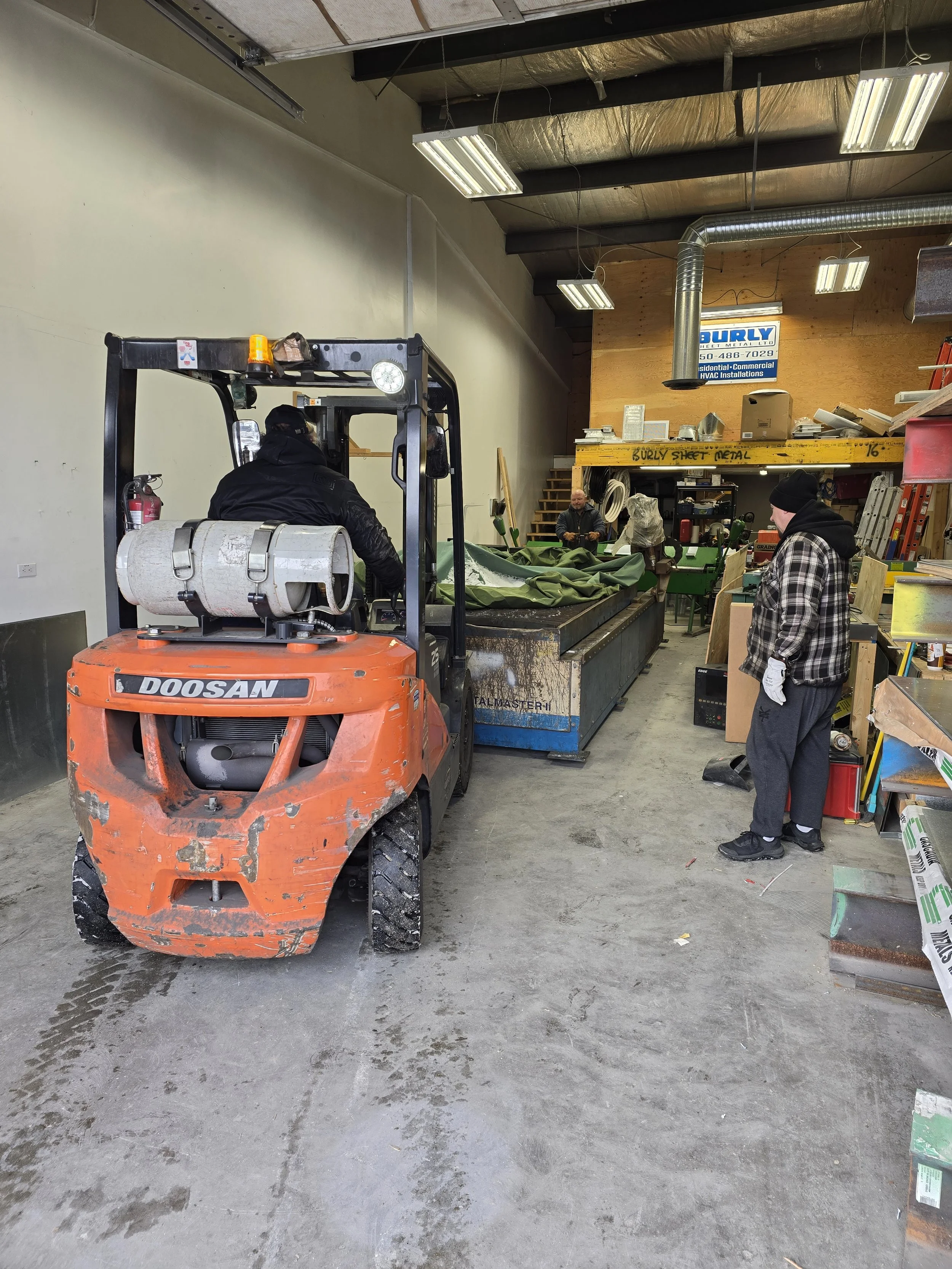 Indoor workshop with three people working at a large worktable, a forklift with a propane tank in foreground, wood and metal supplies around, bright lighting, and a yellow shelf labeled 'Surly Sheet Metal'.