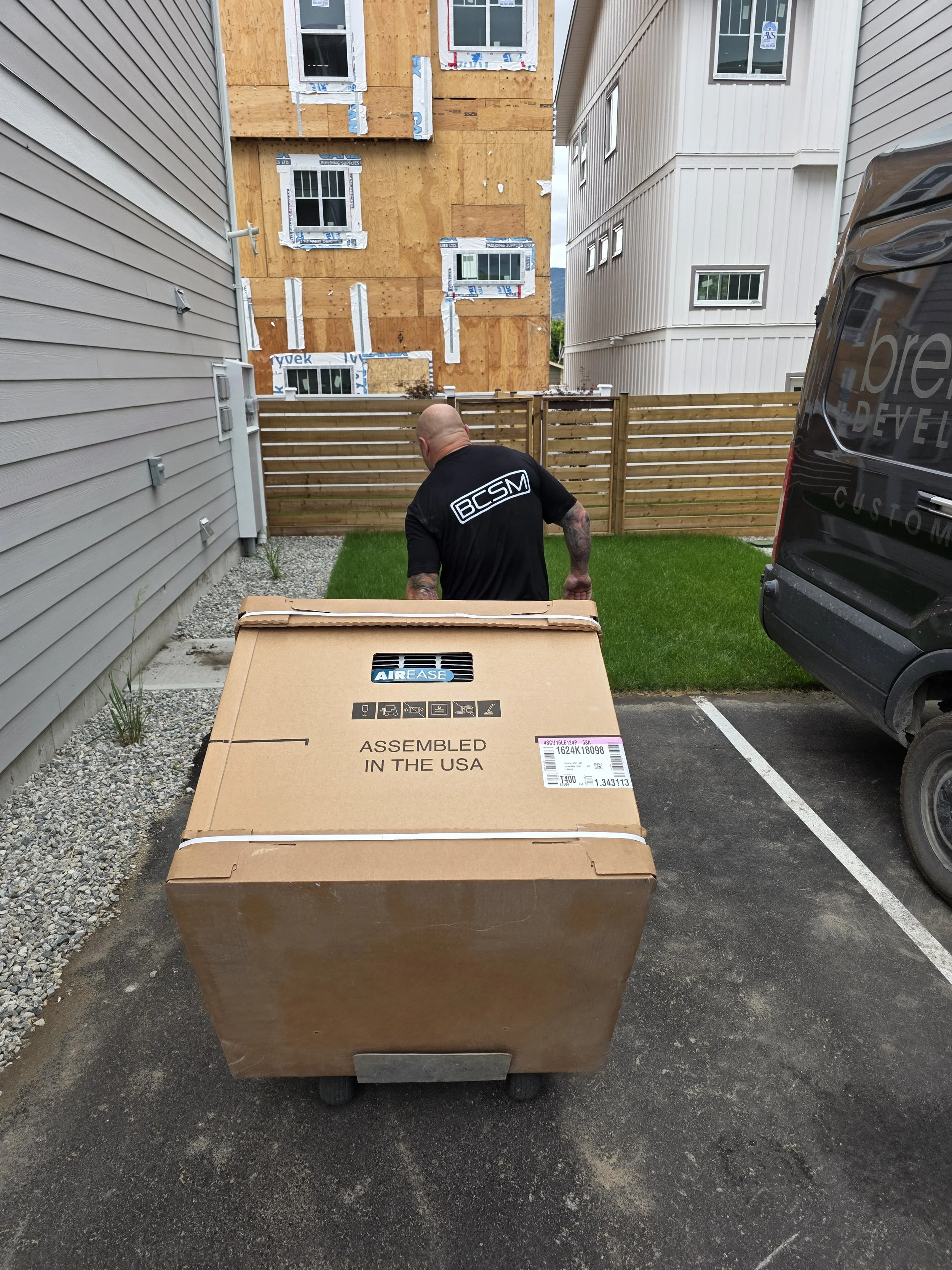 A person with tattoos wearing a black shirt with the letters 'BCSM' on back, seen from behind, pushing a large sealed cardboard box labeled 'Assembled in the USA' and 'Airease' across a parking lot toward a fenced backyard with new construction in th