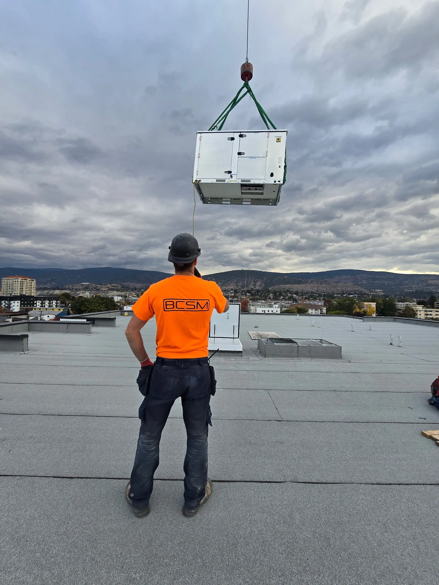 A worker on a rooftop wearing an orange shirt and black helmet observes a large HVAC unit being hoisted by a crane against a cloudy sky with distant mountains and cityscape in the background.