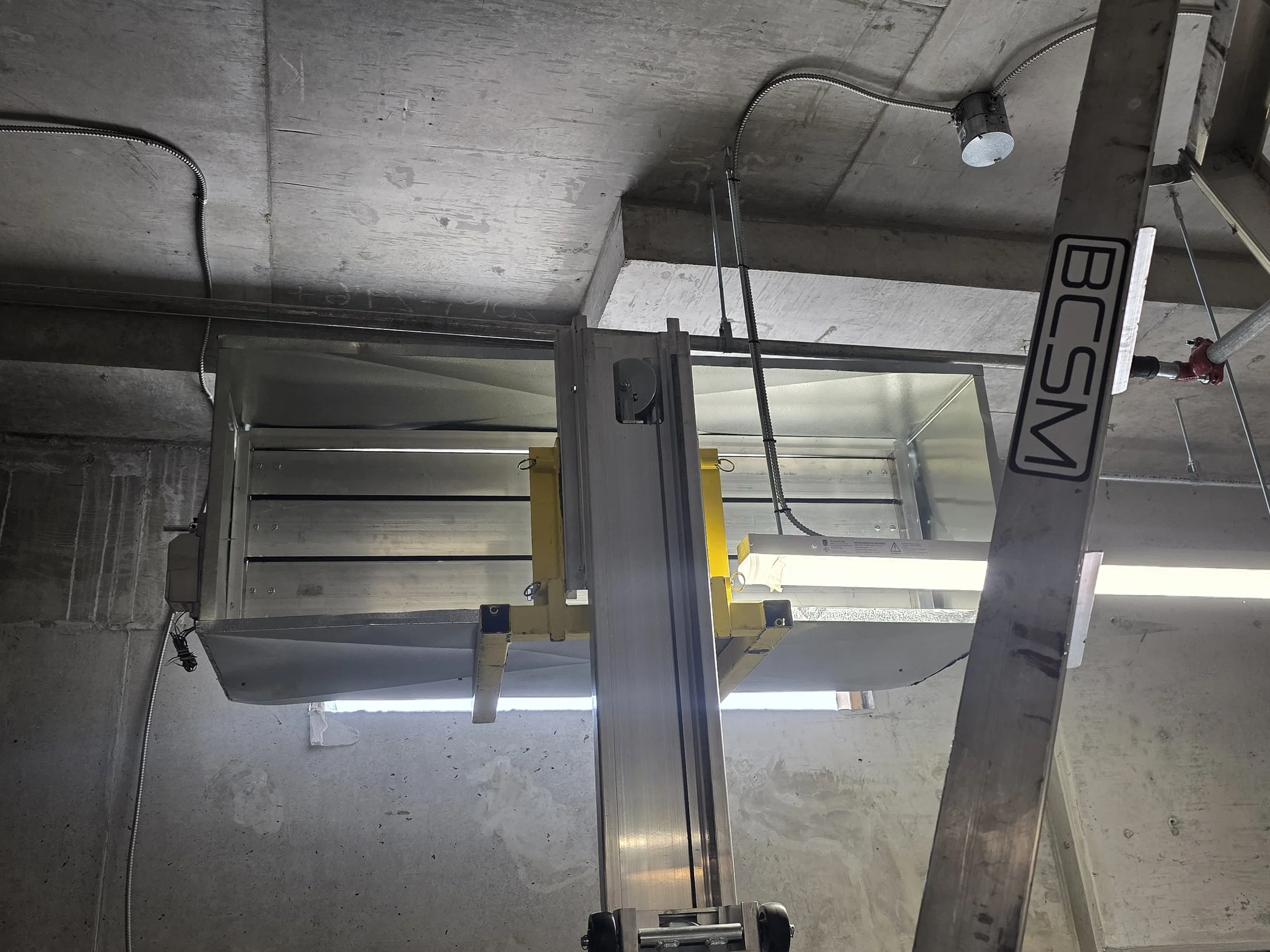 Construction site with exposed concrete walls, electrical conduit, and a sleek metal machine or enclosure, possibly part of HVAC or industrial equipment, with a yellow safety feature and a fluorescent light tube.