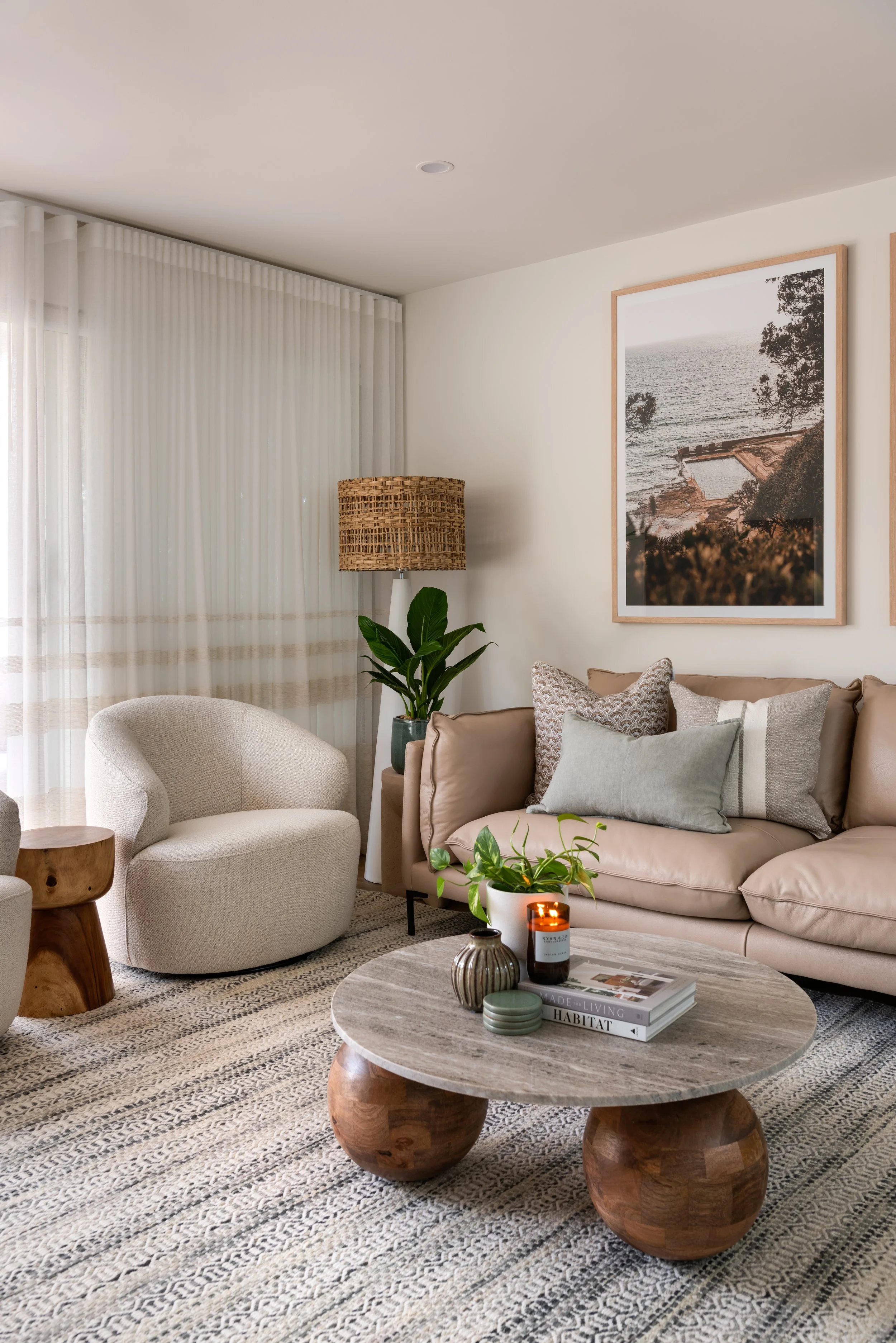 Living room with neutral-toned furniture, a round wooden coffee table with decorative items, a beige sofa with pillows, a white armchair, a green plant, a tall floor lamp with a woven shade, a framed nature photograph on the wall, and sheer curtains covering windows.
