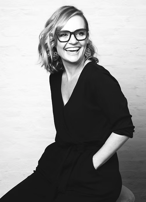 A smiling woman with shoulder-length wavy hair wearing glasses and a black outfit, sitting against a plain light-colored background.
