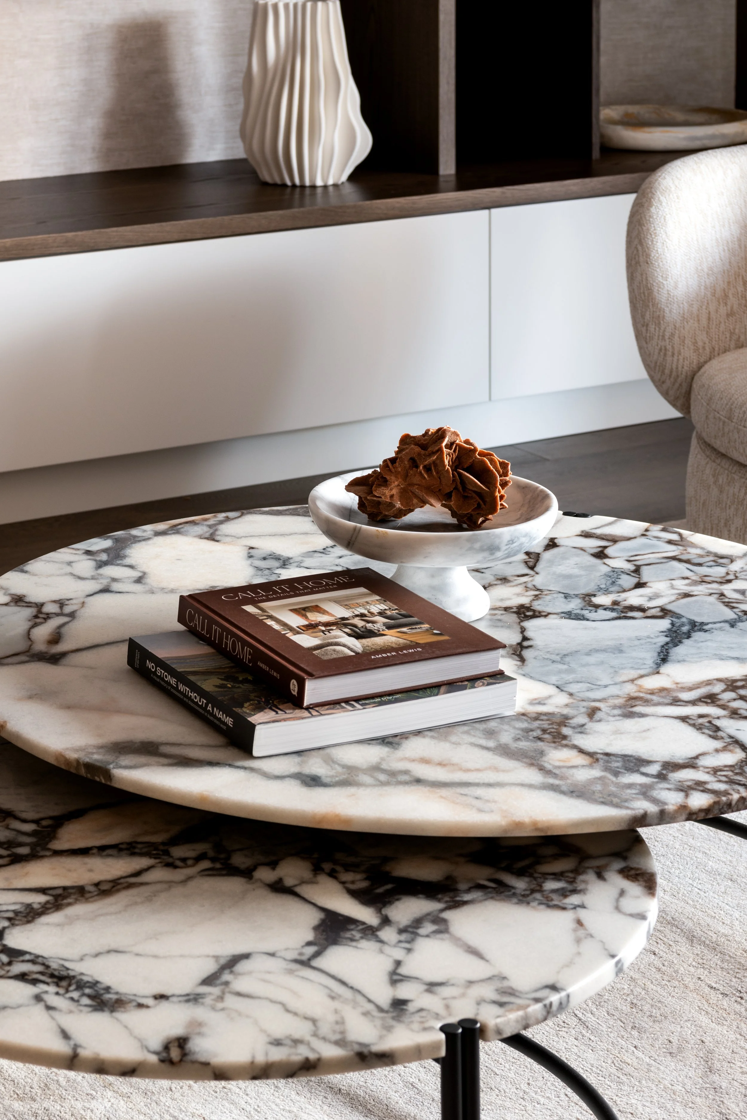 A marble coffee table with books and a decorative bowl on top, in a modern living room with a beige couch and a white cabinet in the background.