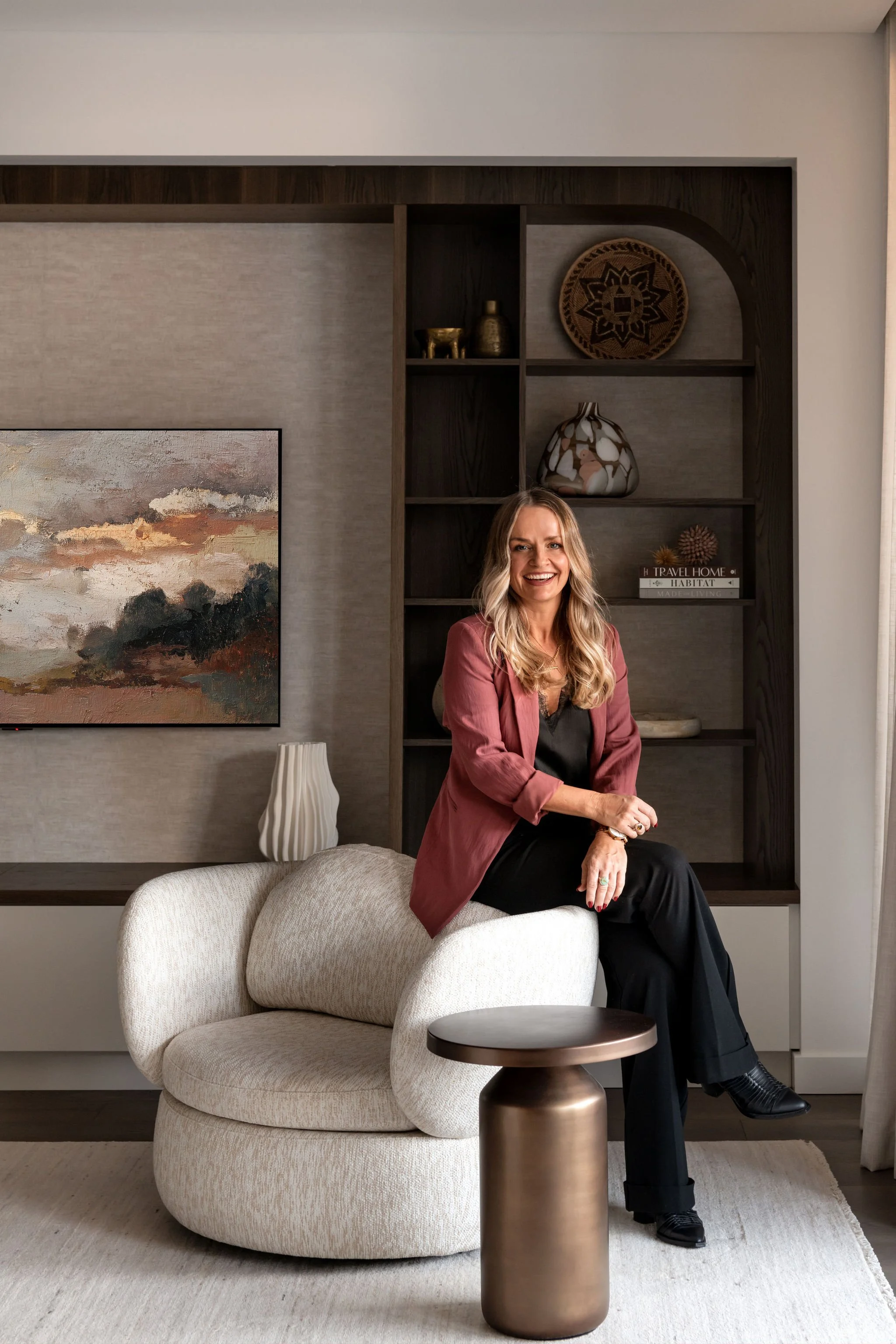A woman with blonde hair, wearing a dark top and a mauve blazer, sitting on a white, modern armchair in a contemporary living room with decorative shelves and artworks, smiling at the camera.