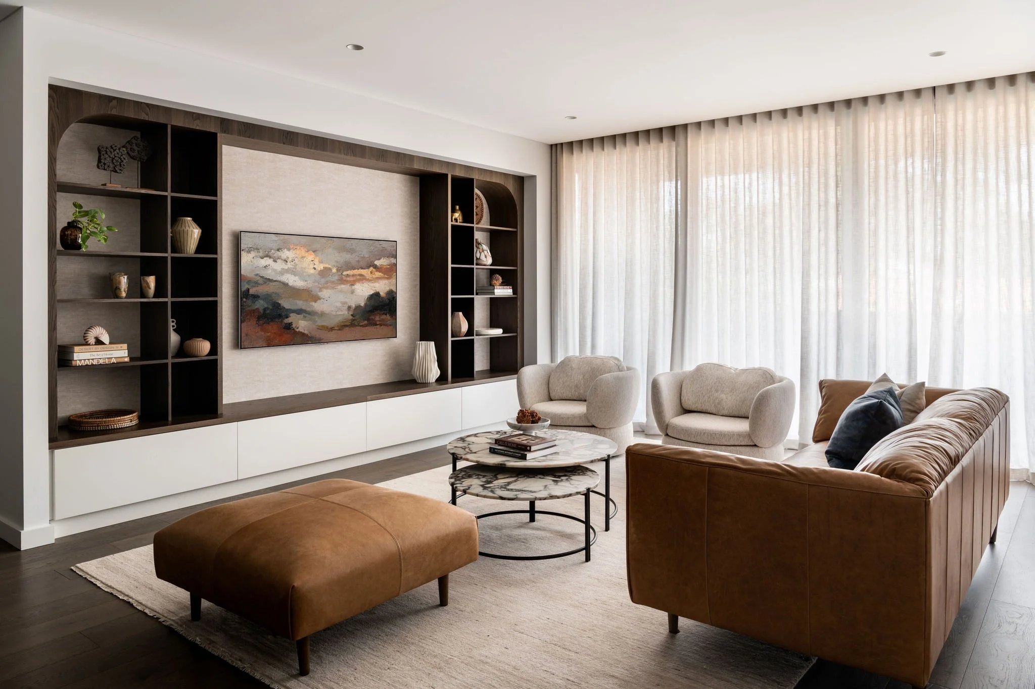 Modern living room with a beige leather sofa, two beige armchairs, a marble-topped coffee table, and built-in shelving unit with decorative items, against a large window with sheer curtains.