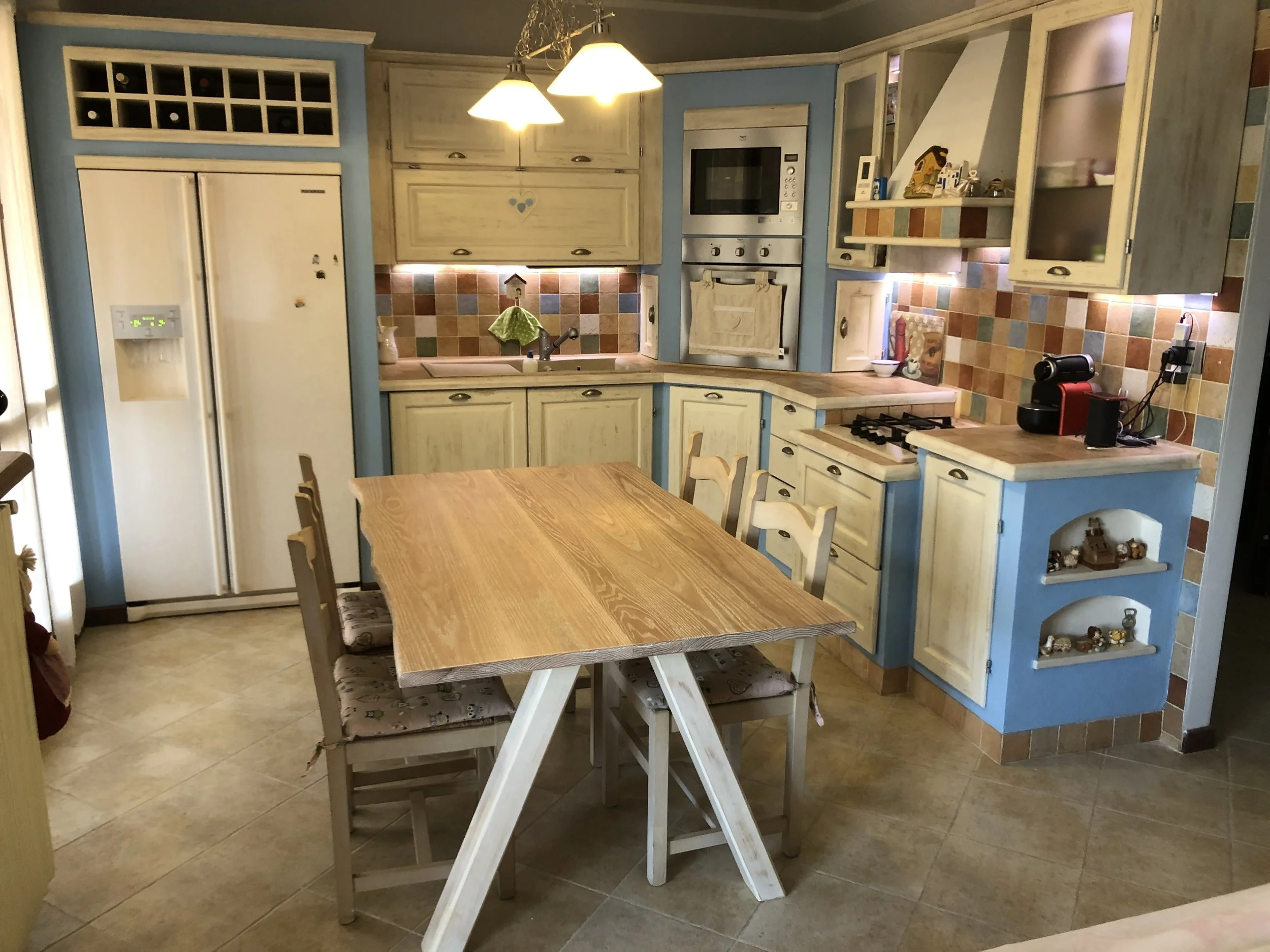 Cucina rustica con mobili chiari e dettagli colorati, tavolo in legno con sedie, elettrodomestici e decorazioni.