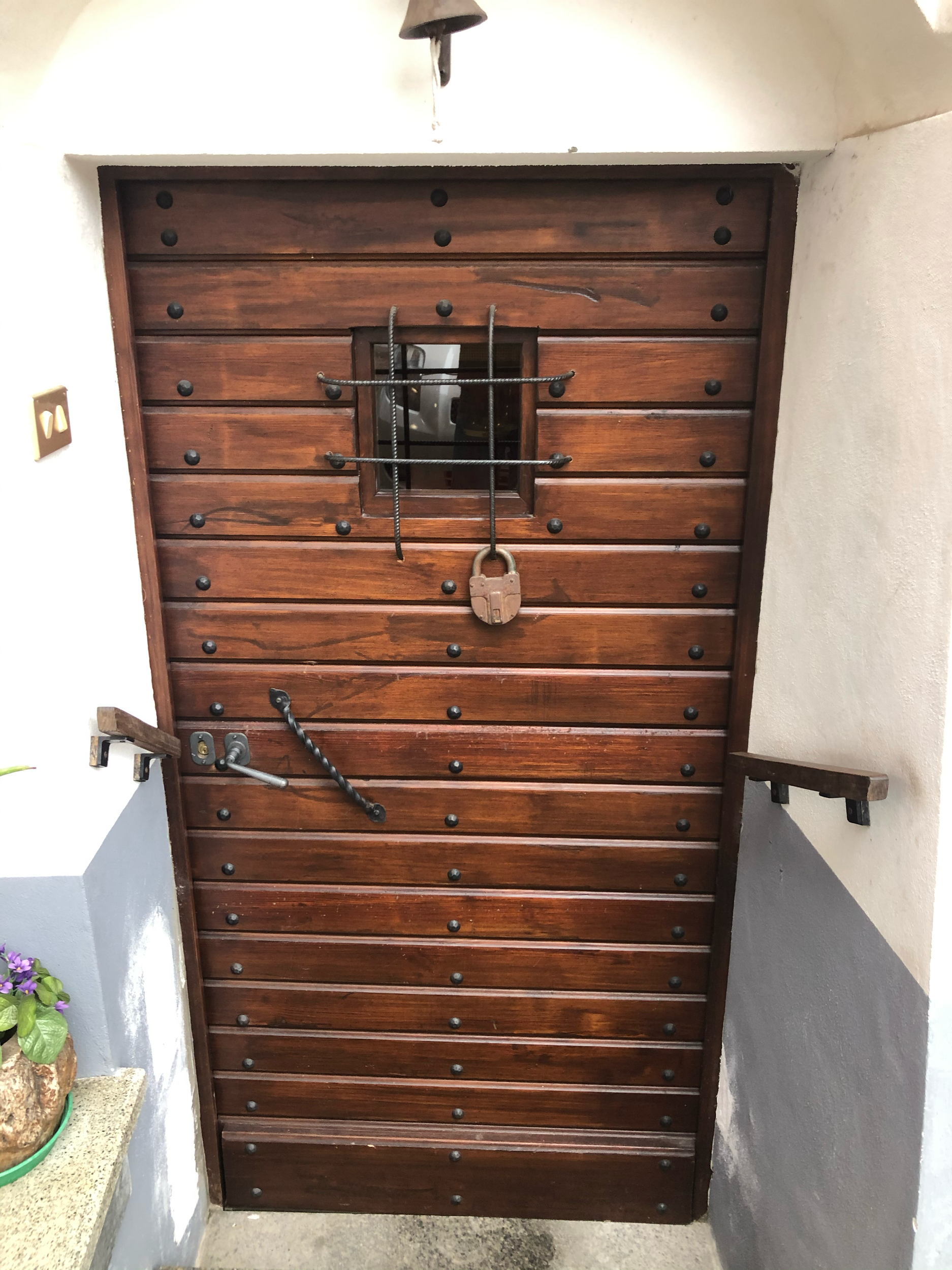 Porta in legno con dettaglio di una piccola finestra, accessori in metallo e una maniglia, decorata con chiodi e inserti metallici, situata all'ingresso di una casa o uno stabile.