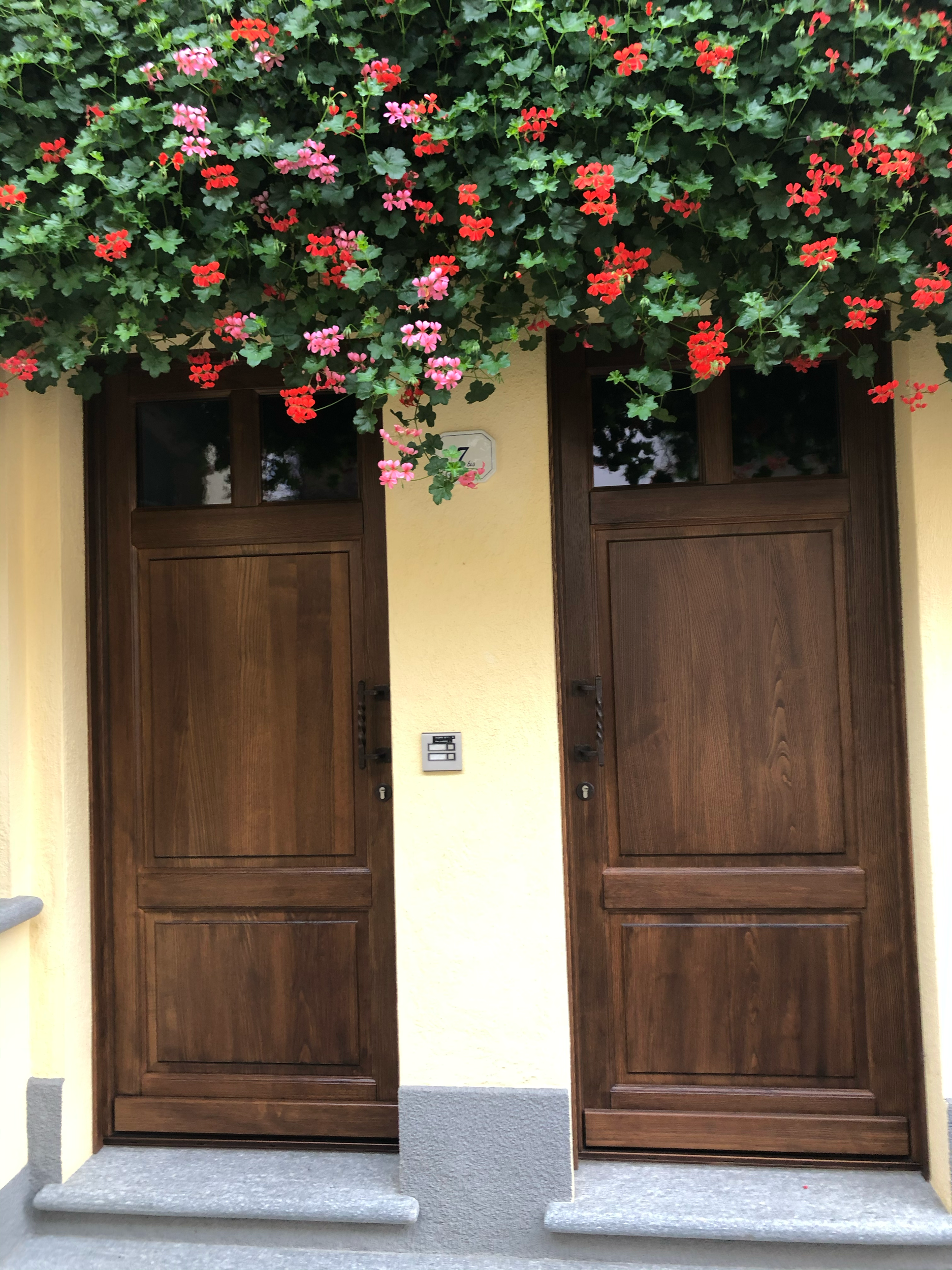 Porta d'ingresso in legno con fianco di fiori rossi e rosa, muro giallo e marciapiede grigio.