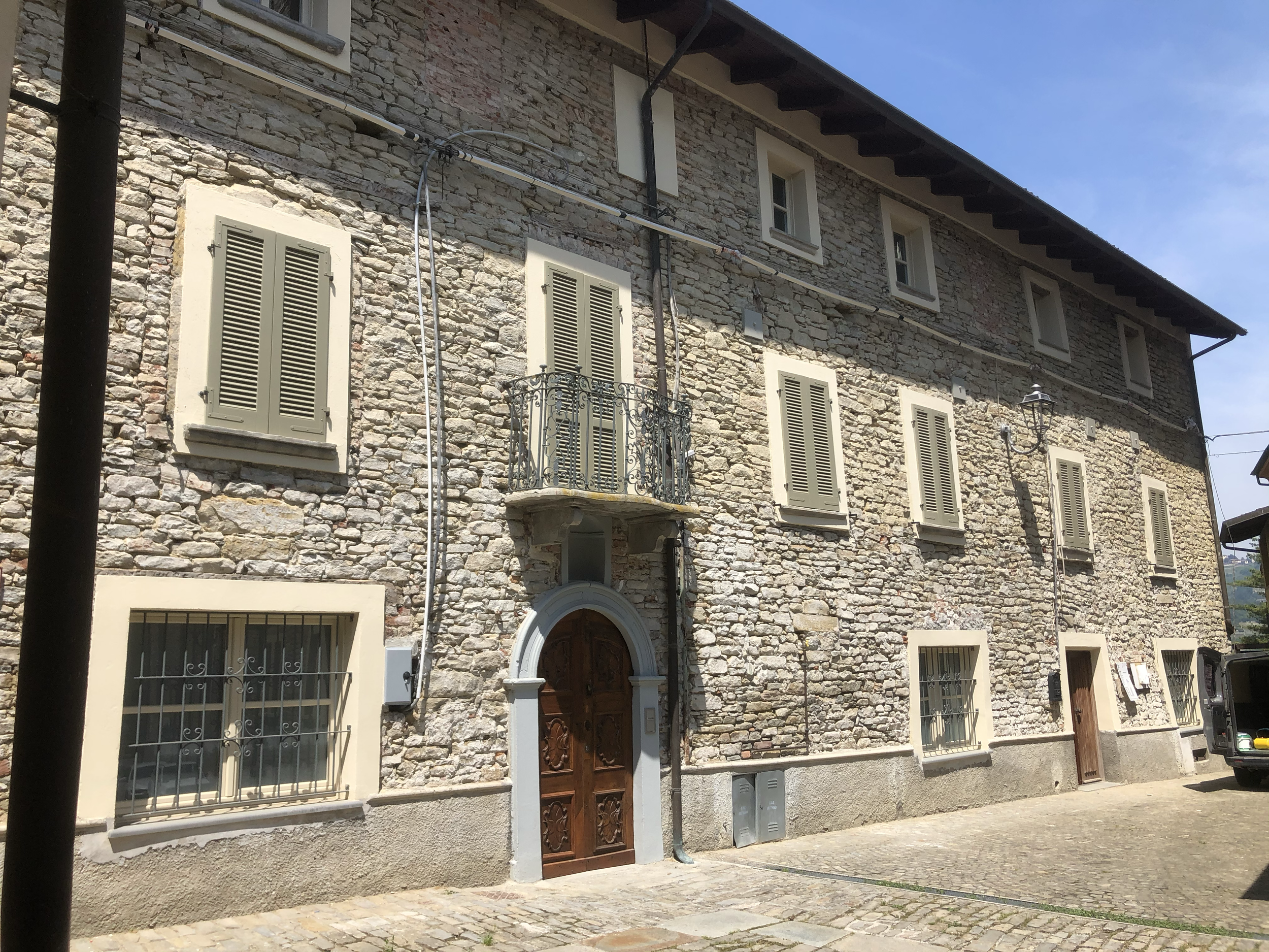 Edificio in pietra con finestre con persiane verdi e porta in legno decorata, balcone con ringhiera in ferro battuto sopra la porta, strada in pietra davanti, lampione e cablaggi visibili.