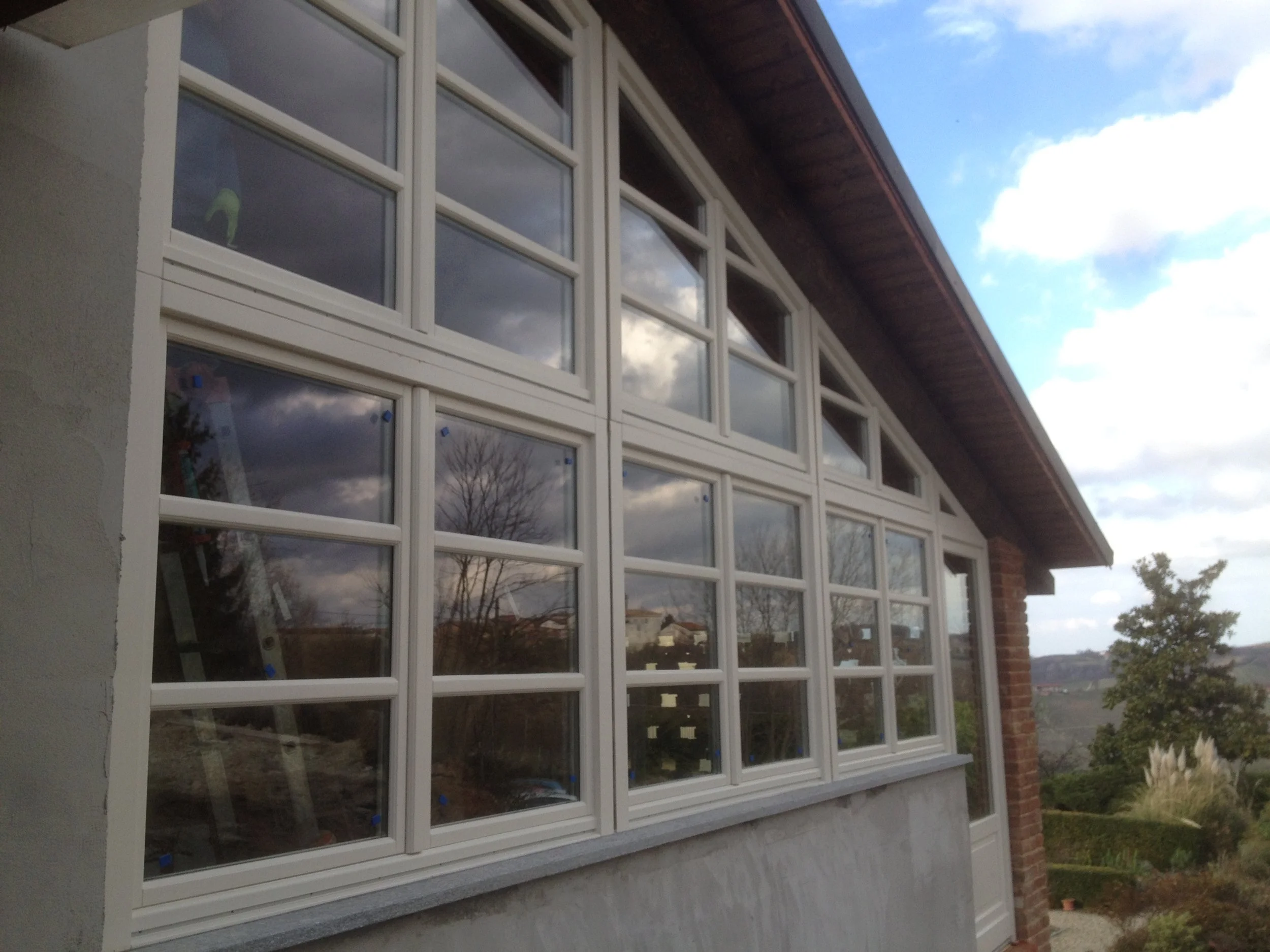 Finestra di una casa con vetri e strutture bianche che riflettono il cielo nuvoloso e il paesaggio circostante, con alberi e colline sullo sfondo.