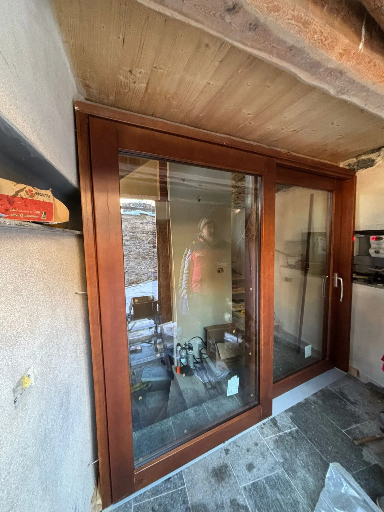 Una porta scorrevole in legno con vetri, installata in un ambiente casalingo o commerciale, con strumenti e materiali sul pavimento e riflessi di persone nello specchio.