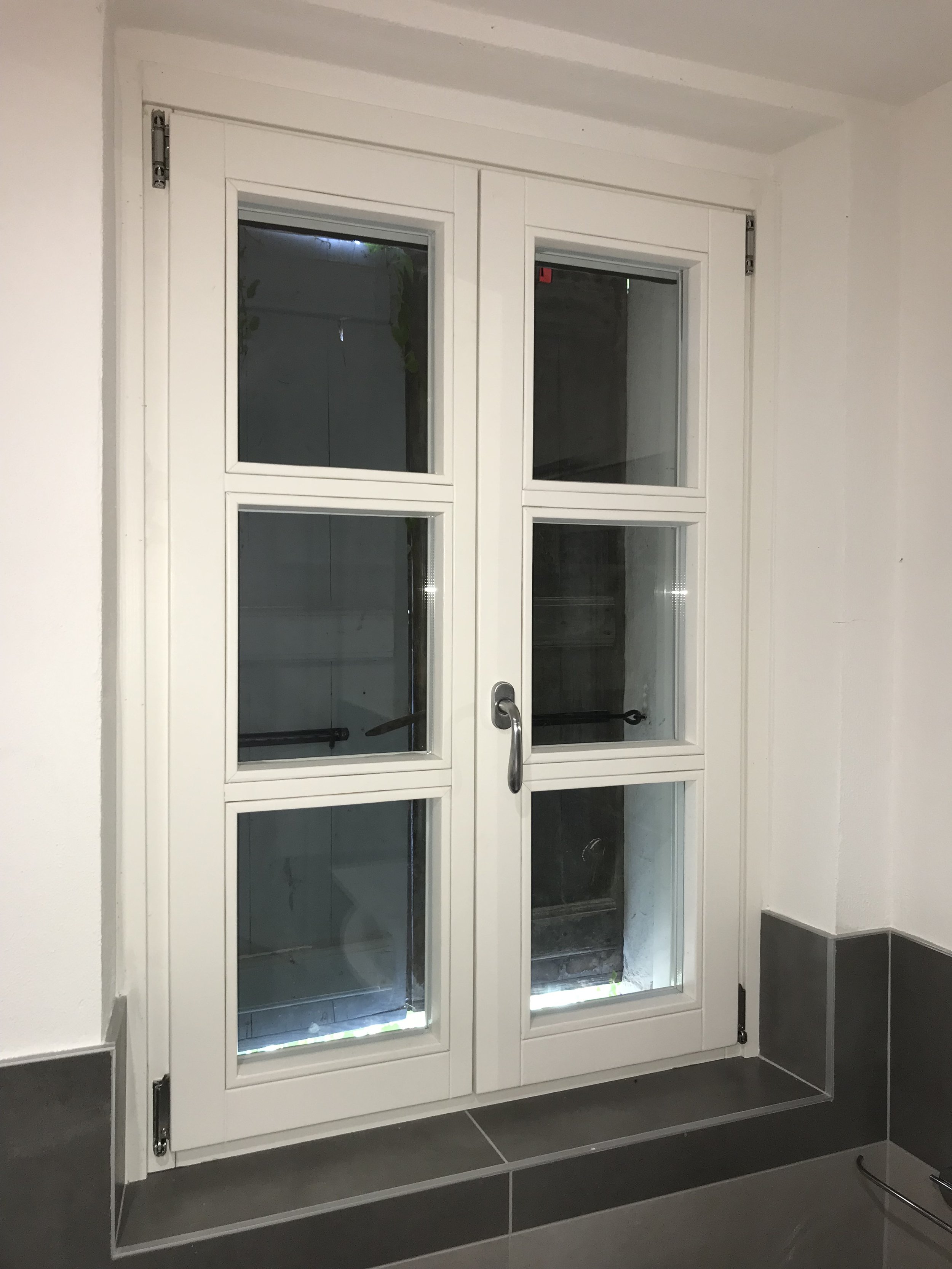 Finestra bianca con quattro pannelli di vetro in una stanza con pareti bianche e piastrelle grigie.