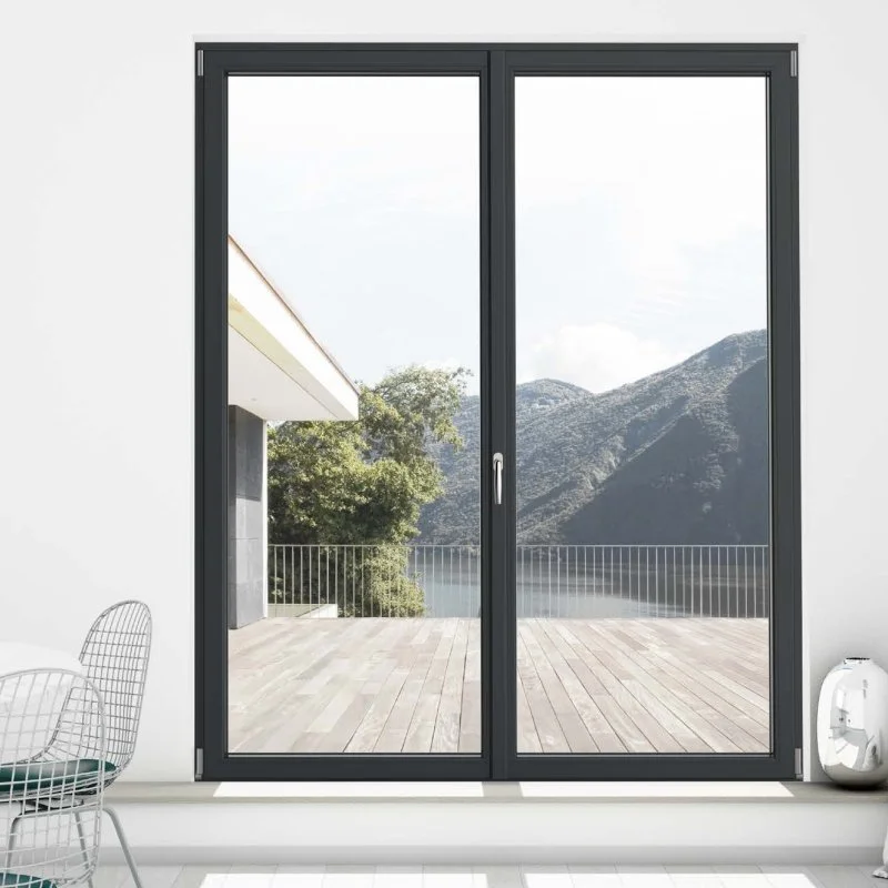 Foto di una porta finestra a doppio vetro con telaio nero, che si apre su un terrazzo con vista sulle montagne e un albero. L'interno della stanza è minimalista, con una sedia in metallo bianco e un oggetto bianco a forma di parallelepipedo sul pavimento.