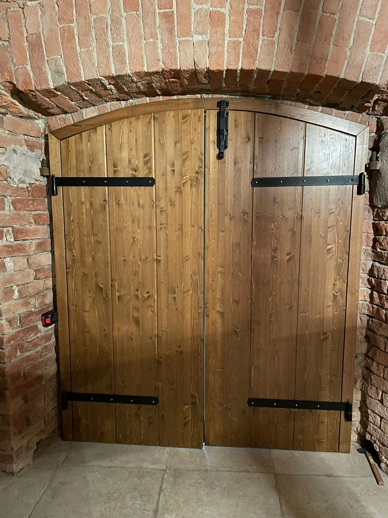 Doppia porta in legno con cerniere nere, installata tra due pareti di mattoni, sul pavimento di piastrelle.