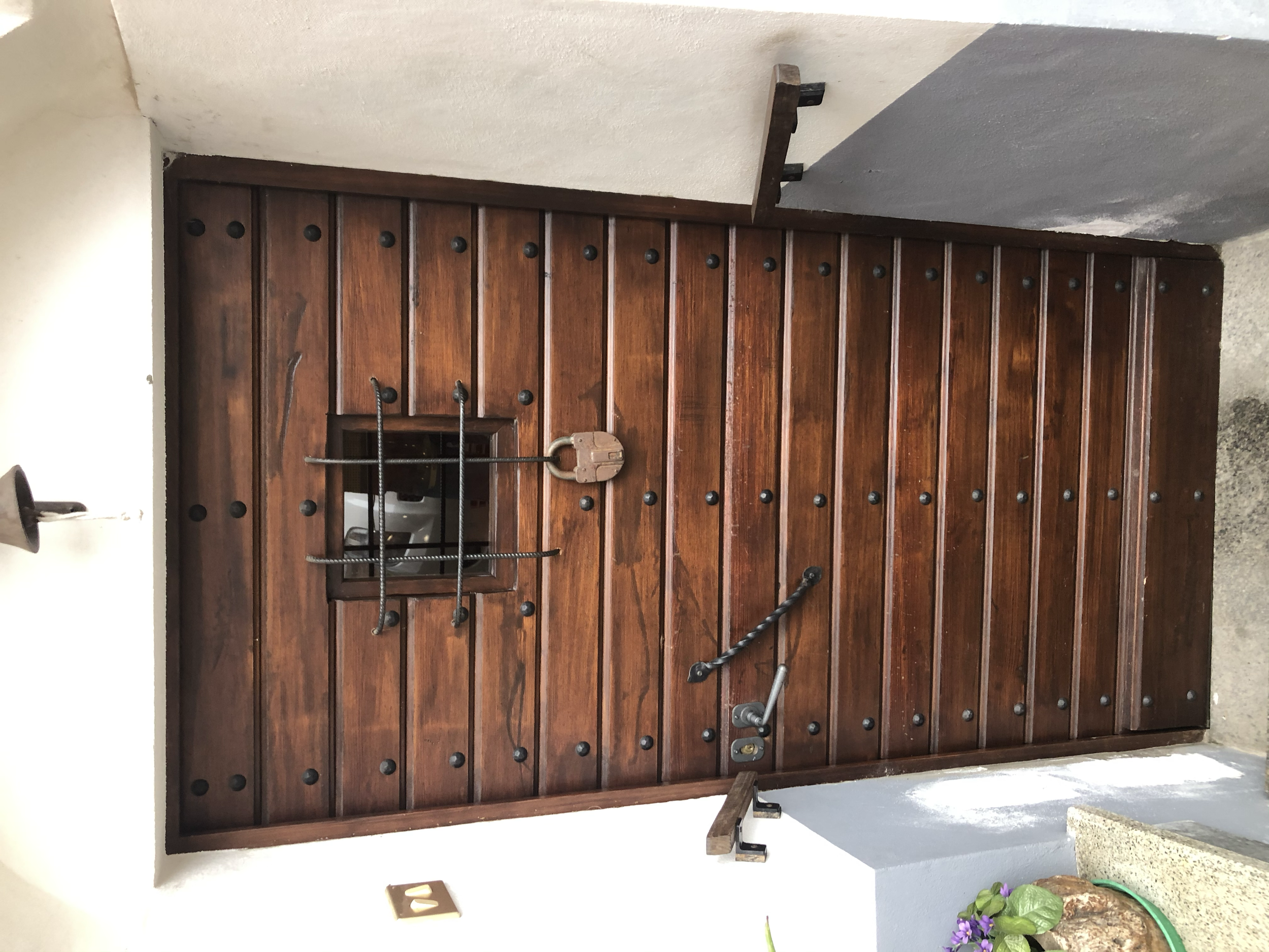Porta in legno con dettaglio di una piccola finestra, accessori in metallo e una maniglia, decorata con chiodi e inserti metallici, situata all'ingresso di una casa o uno stabile.
