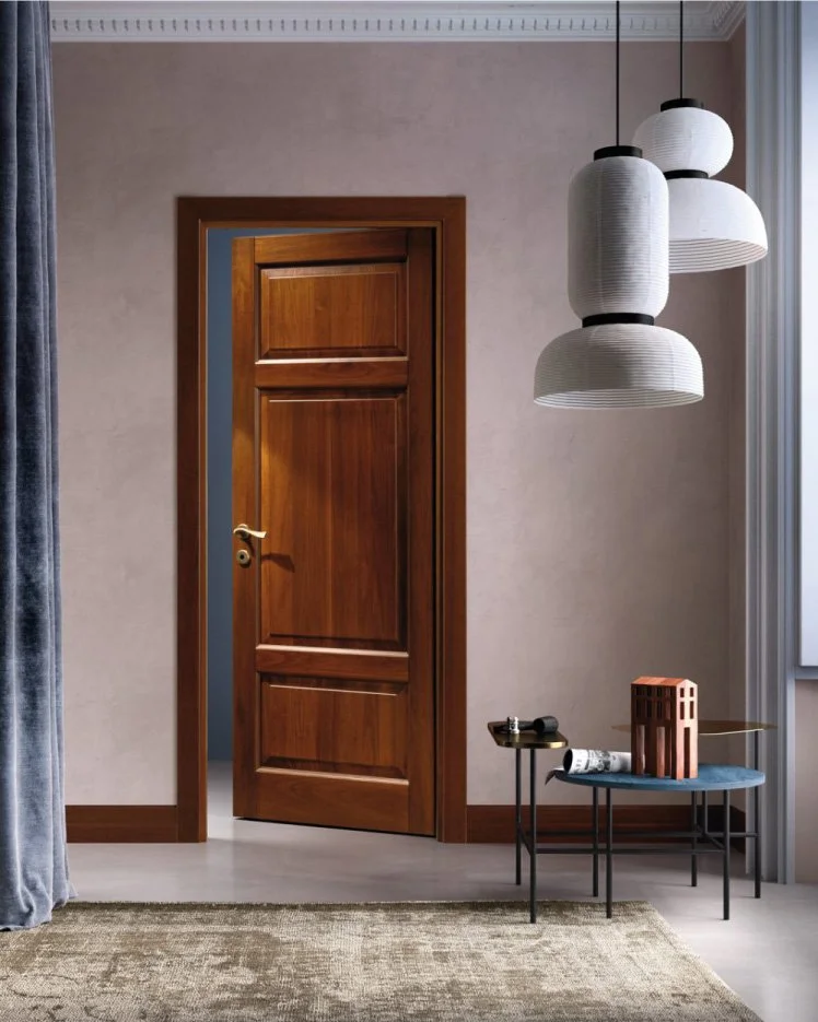 Una stanza con pareti beige, porta in legno chiaro leggermente aperta, lampade pendenti bianche, due tavolini neri con oggetti decorativi e un tappeto beige.