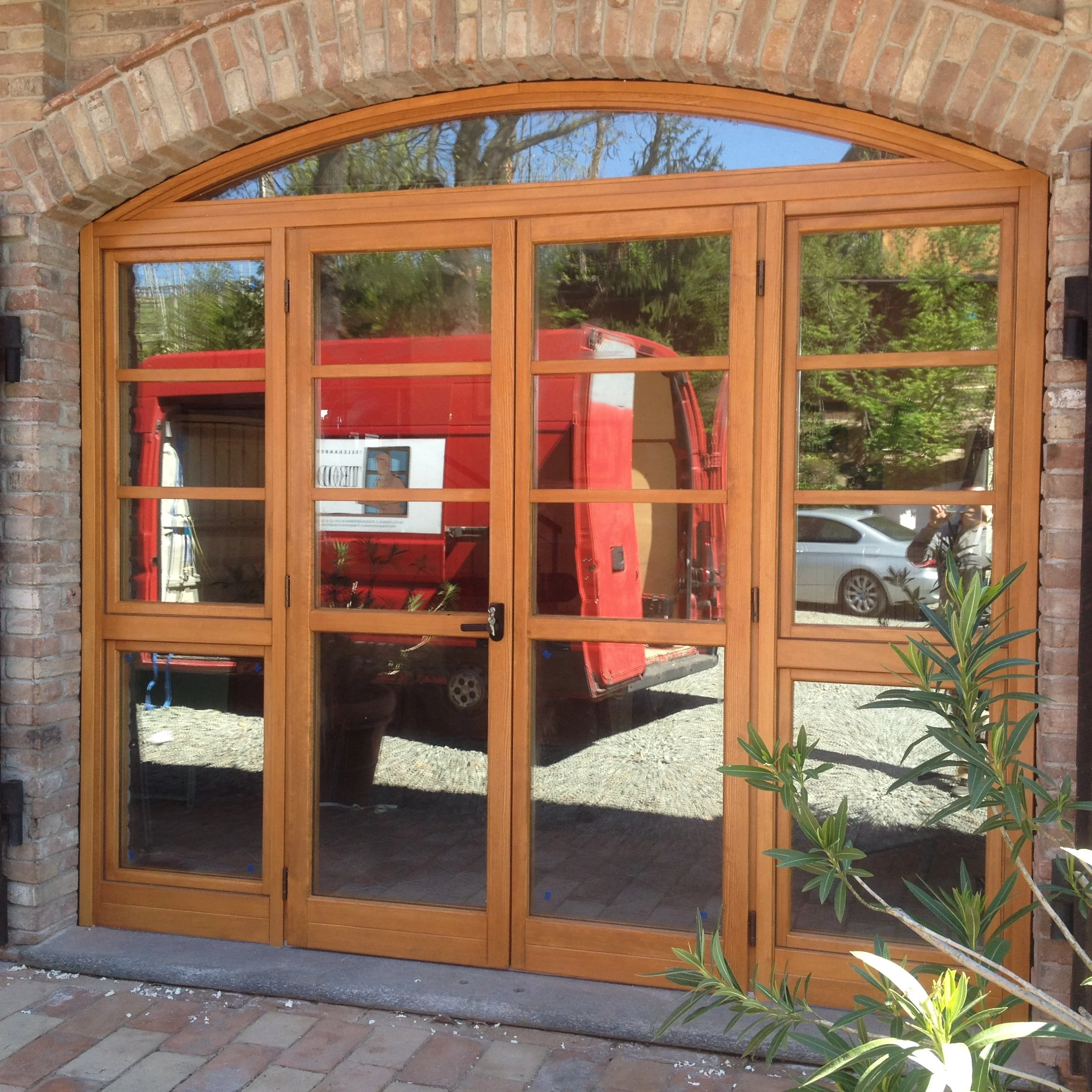 Porta scorrevole in legno con vetri, inserita in un arco in mattoni, con piante verdi sul lato destro.