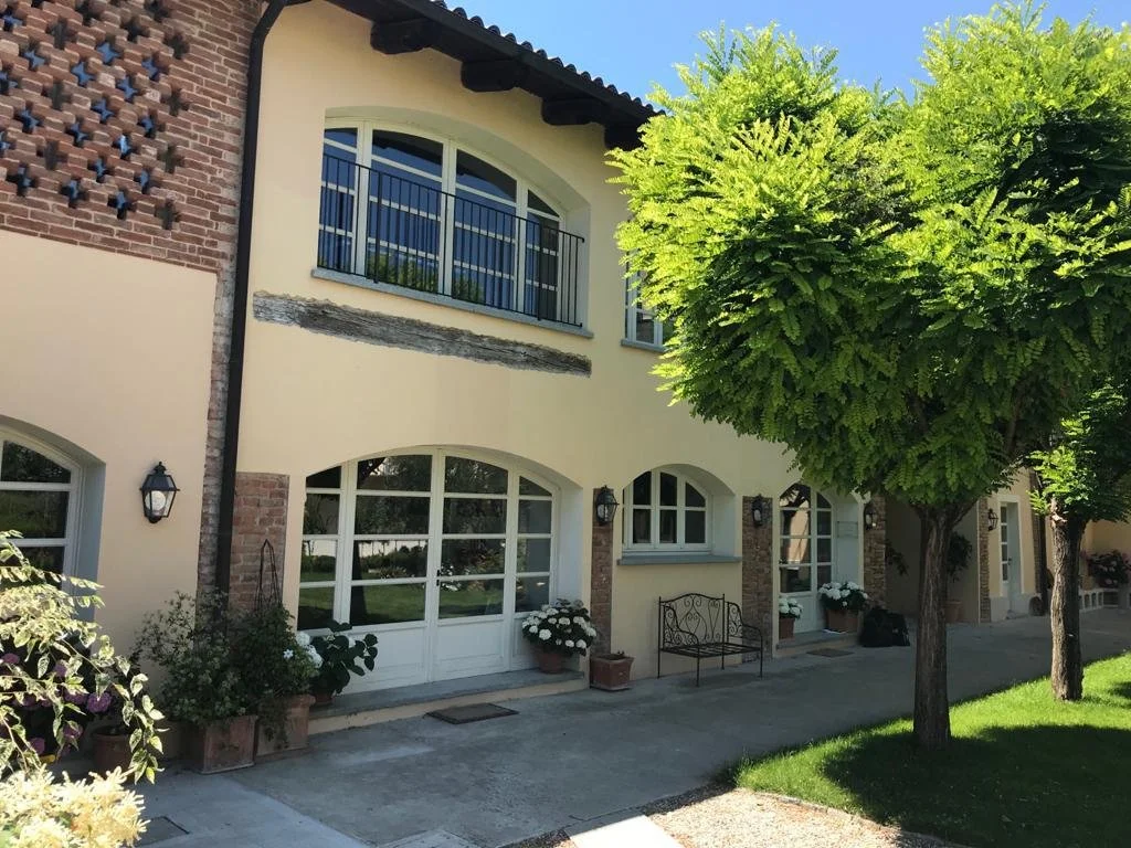 Casa con facciata in stile rurale, con grandi finestre ad arco, balcone al primo piano con ringhiera di metallo, pareti chiare e dettagli in mattoni, circondata da alberi e piante decorative.