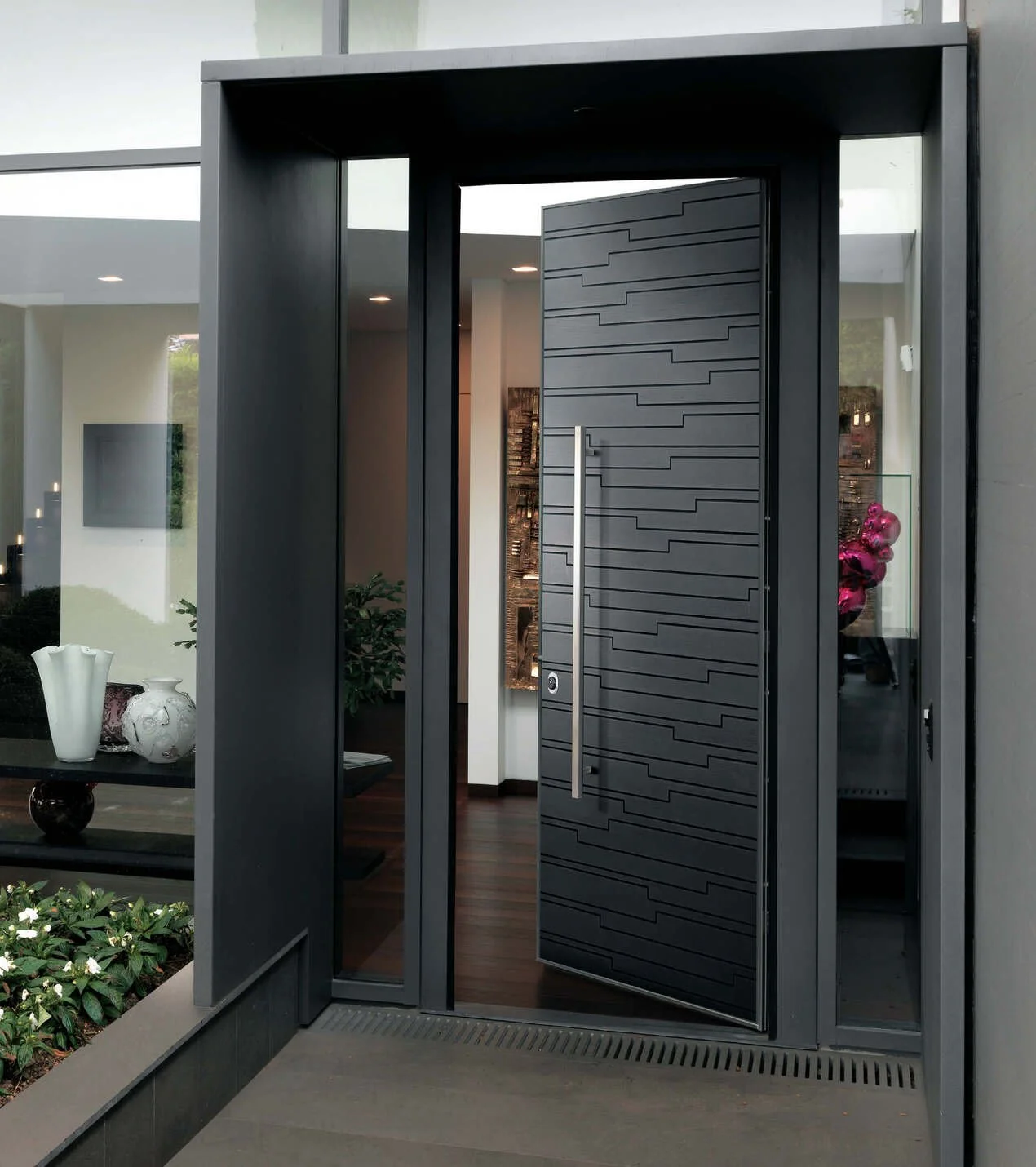 Porta d'ingresso moderna nera con design geometrico, aperta, affacciata su una casa elegante con decorazioni e piante.
