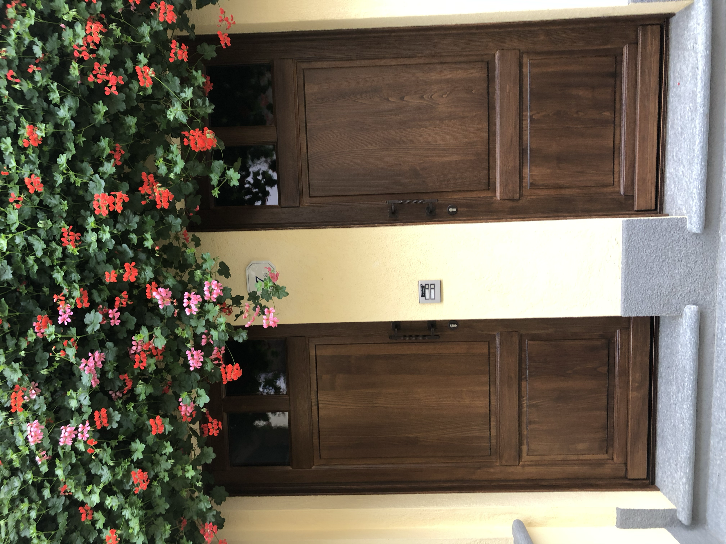 Porta d'ingresso in legno con fianco di fiori rossi e rosa, muro giallo e marciapiede grigio.