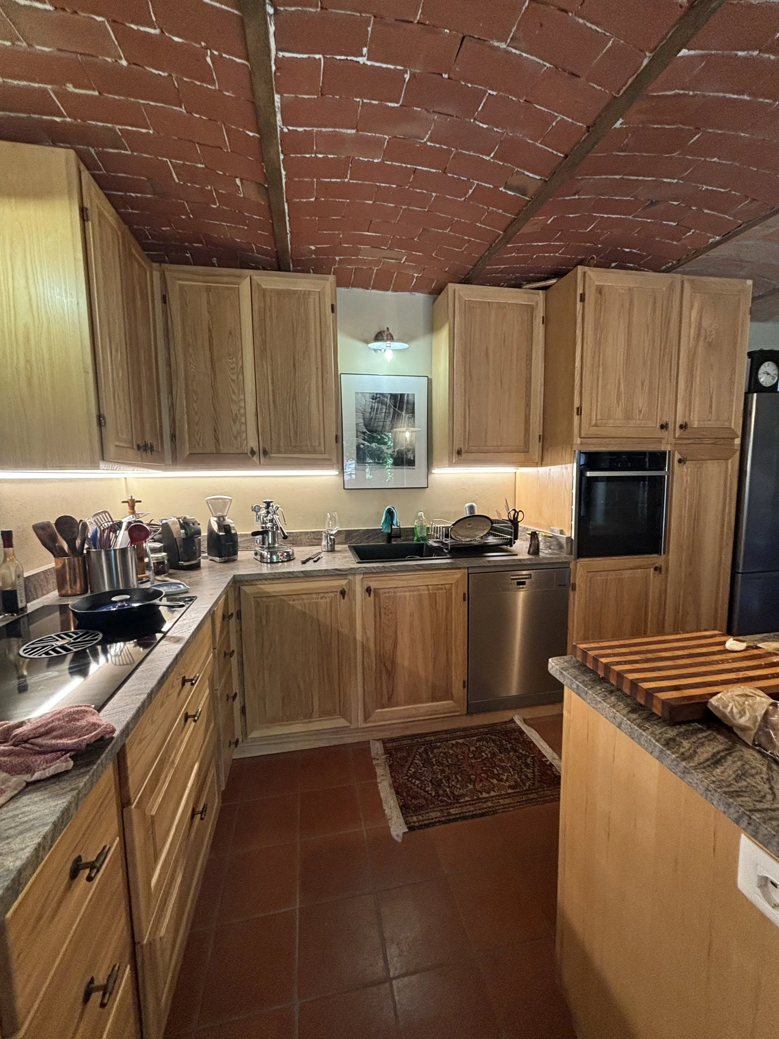 Cucina con armadietti di legno chiaro, piano di lavoro in granito, e un soffitto in mattoni rossi a volta. Sul piano ci sono vari elettrodomestici e utensili da cucina, oltre a un tagliere di legno.