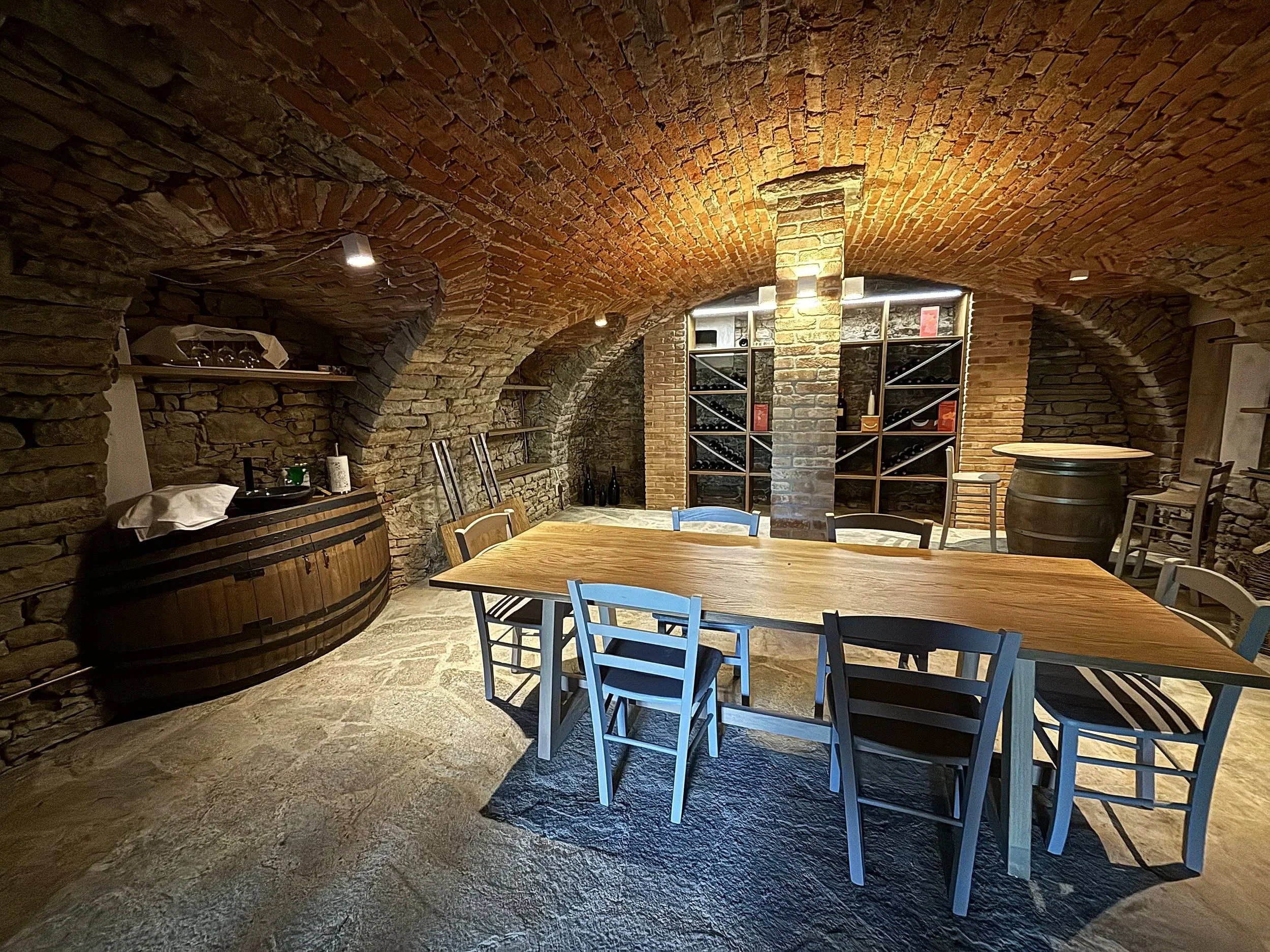 Salone di vinoteca con pareti in pietra, tavolo in legno circondato da sedie in vari colori, scaffali con bottiglie di vino, lampade a soffitto, pavimento in pietra.