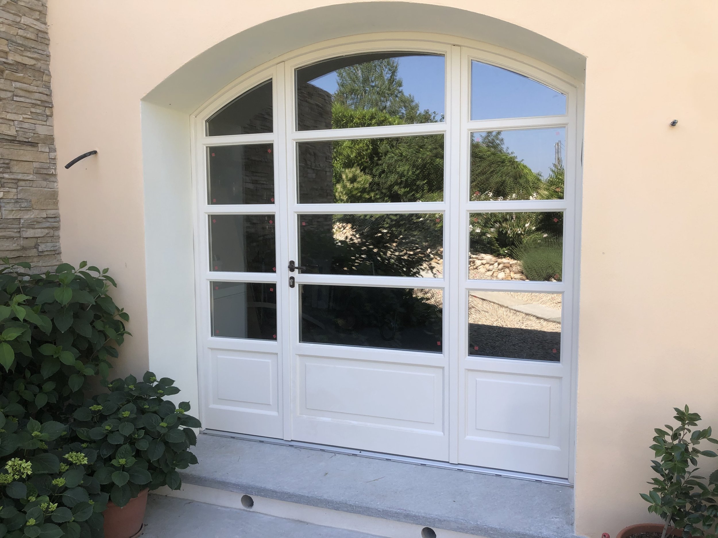 Porta bianca con vetri divisi in più pannelli e arco superiore, ingresso in un cortile con piante verdi ai lati.