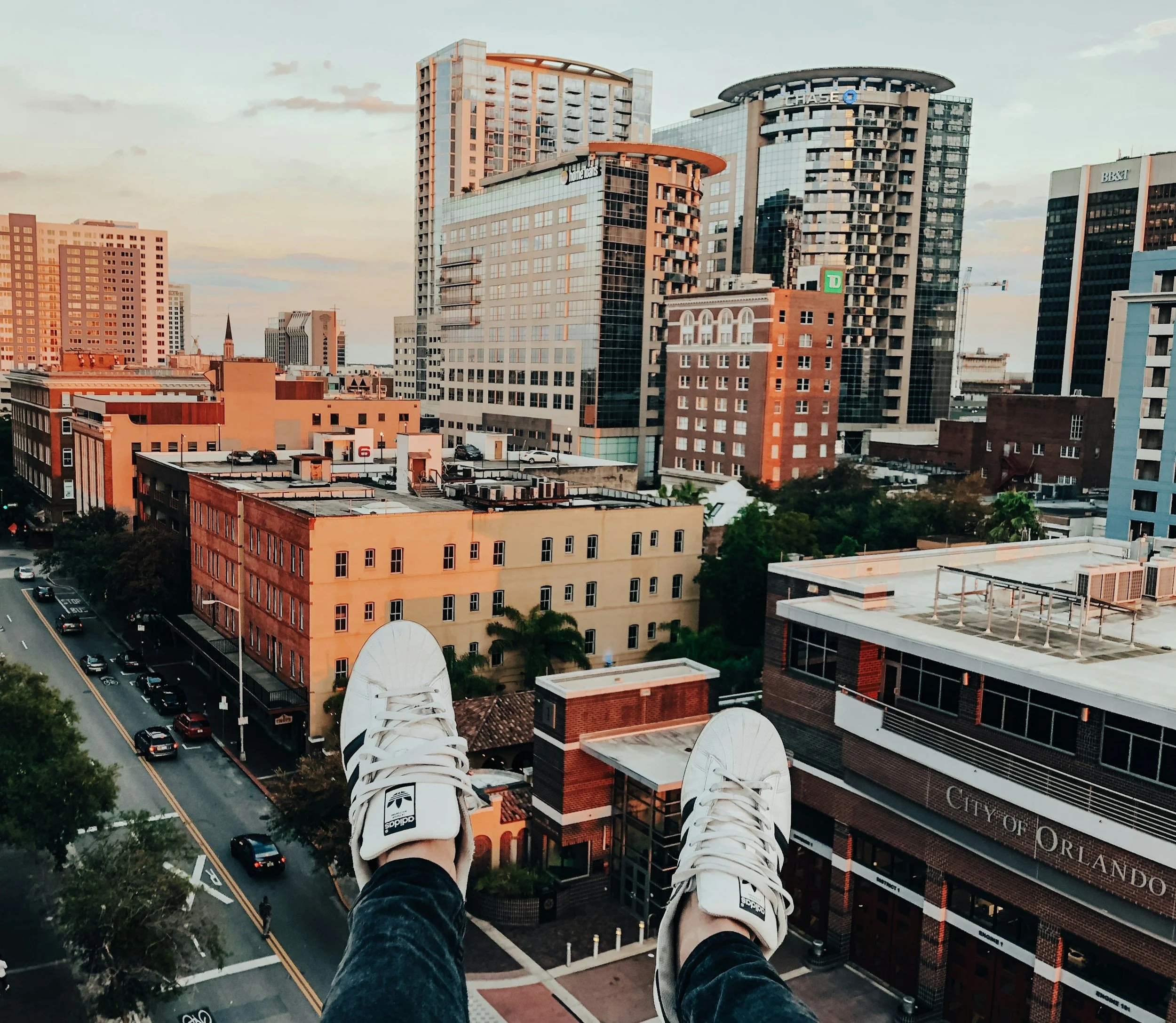 View of a city skyline with various high-rise buildings from a person's perspective, who is sitting on a ledge or rooftop with legs and white sneakers visible in the foreground.
