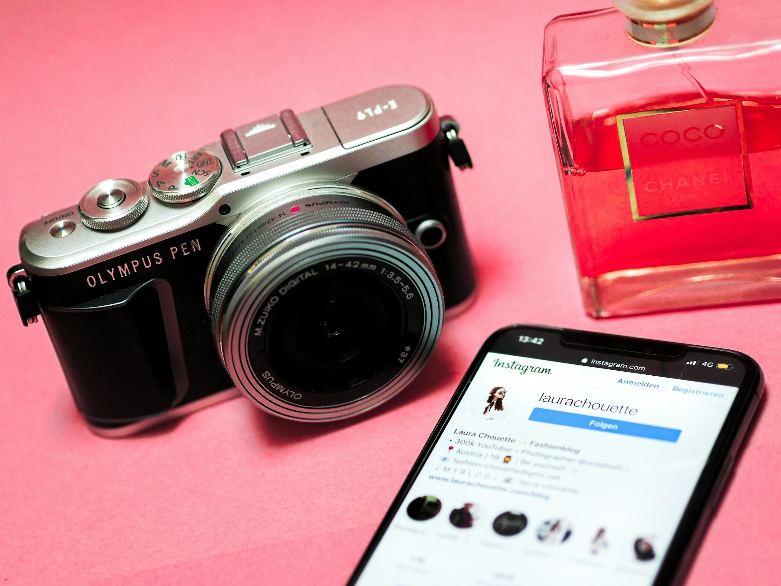 A silver Olympus Pen camera, a bottle of Chanel Coco perfume, and a smartphone displaying an Instagram profile on a pink background.