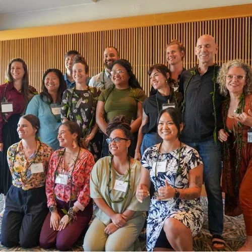 Group of people posing for a photo at an indoor event or conference, smiling and standing against a wooden wall.
