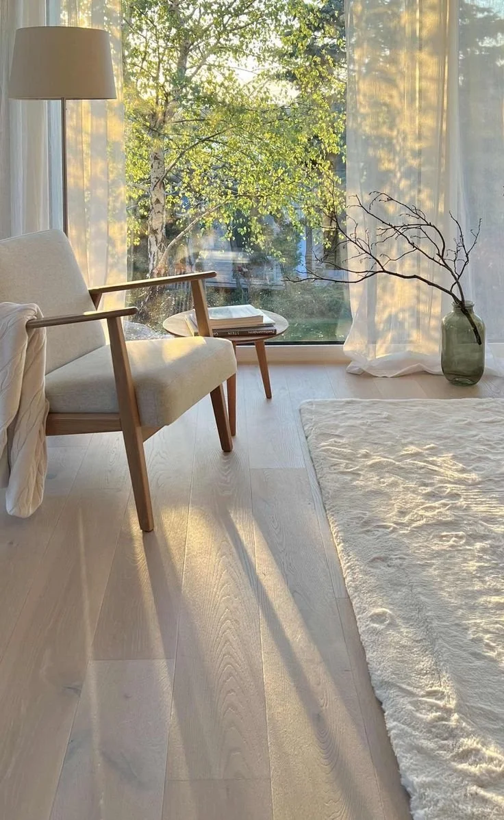 Image of a cozy living space with sunlight streaming through a large window. There is a beige armchair with wooden arms, a small side table holding a couple of books, a floor lamp in the corner, a sheer white curtain, a large glass vase with bare branches, a cream-colored rug, and a view of trees outside.
