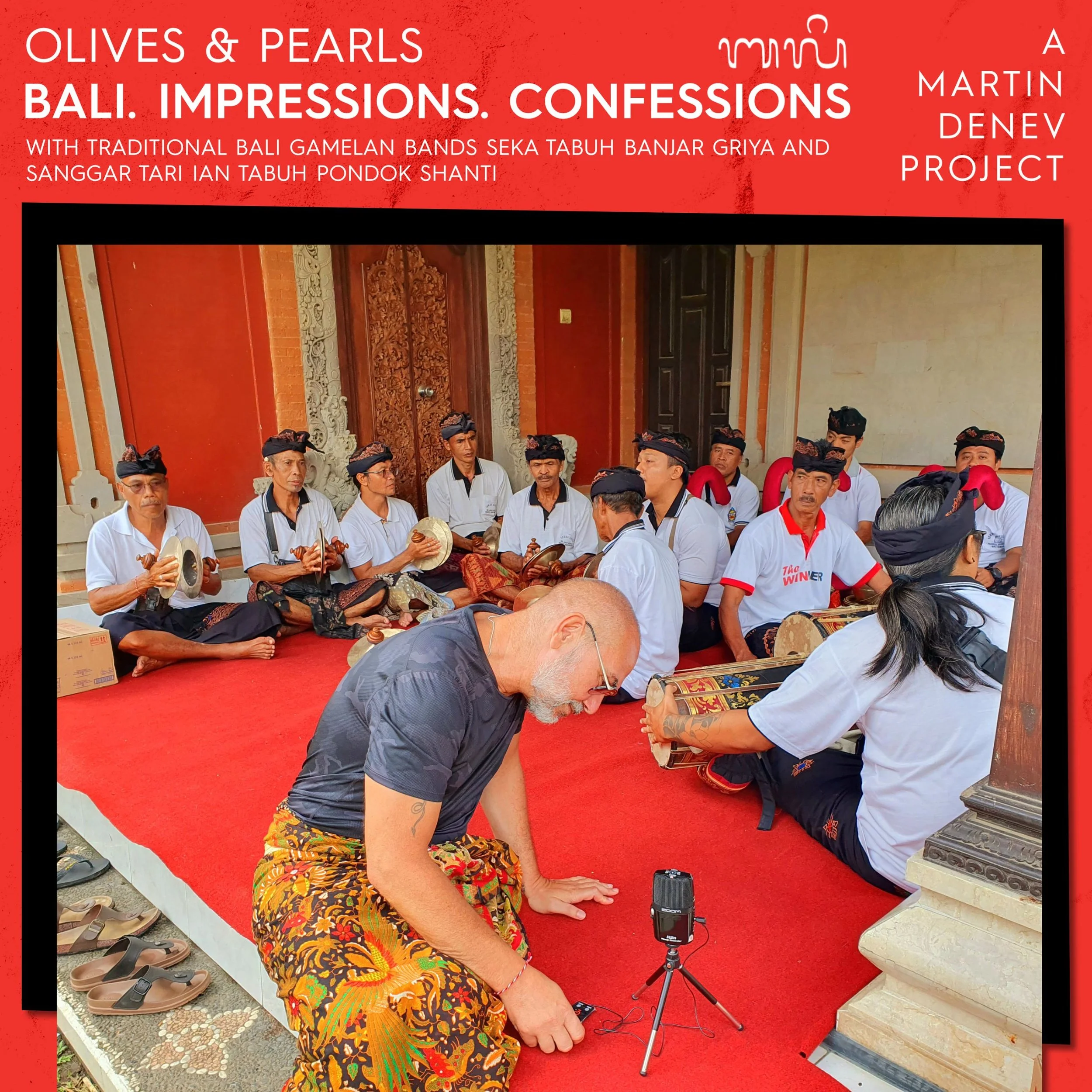 A group of Balinese musicians sitting on a red carpet, playing traditional instruments during a cultural performance, with a man in the foreground kneeling on the carpet, adjusting a small camera on a tripod.
