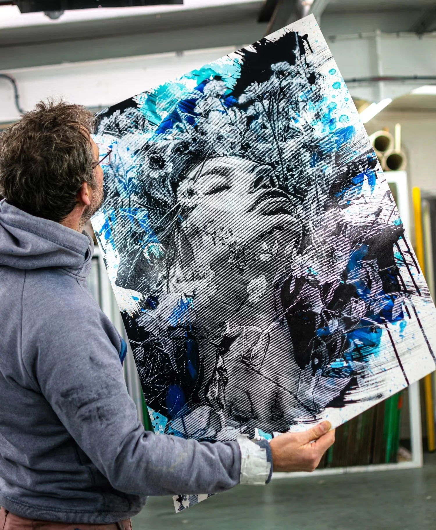 An artist holds a large canvas artwork featuring a stylized female face with floral and nature motifs, in a workspace or studio.