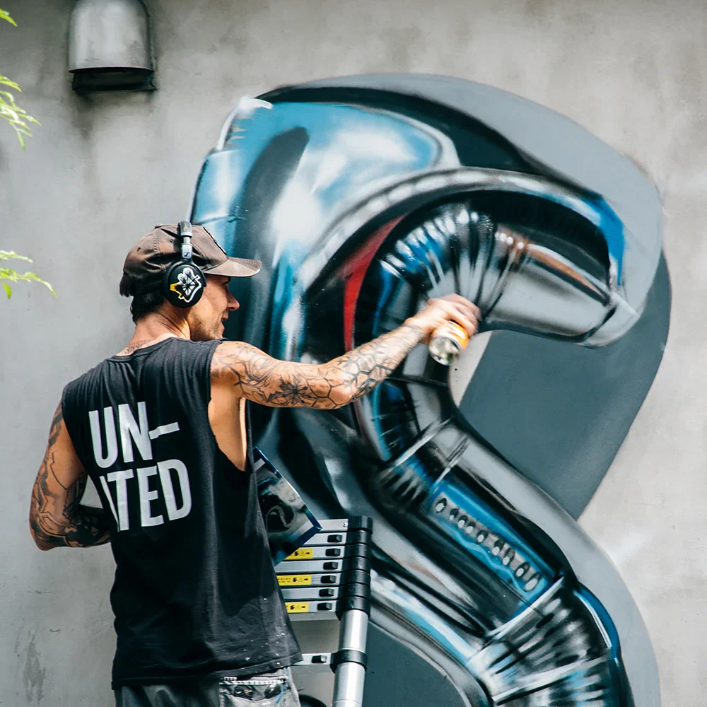 A graffiti artist wearing a black sleeveless shirt with 'United' written on the back, a brown cap, and headphones, spray-painting a detailed mural of a gun on a concrete wall. The artist has tattoos on his arms and is using spray paint cans on a portable cart.