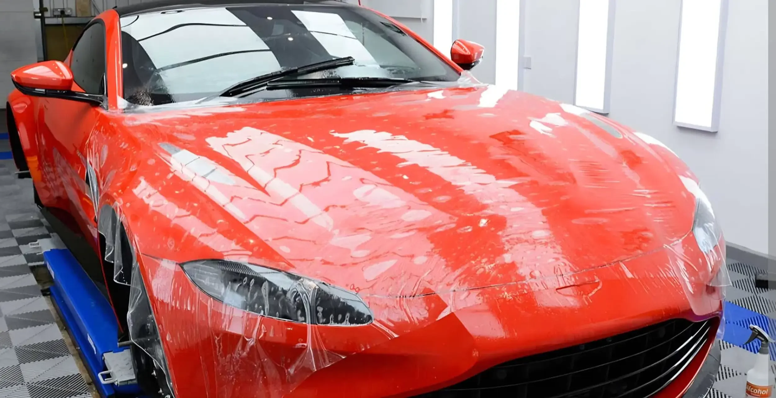 A sleek, red sports car sits indoors on a checkered floor, partially covered in a protective film.