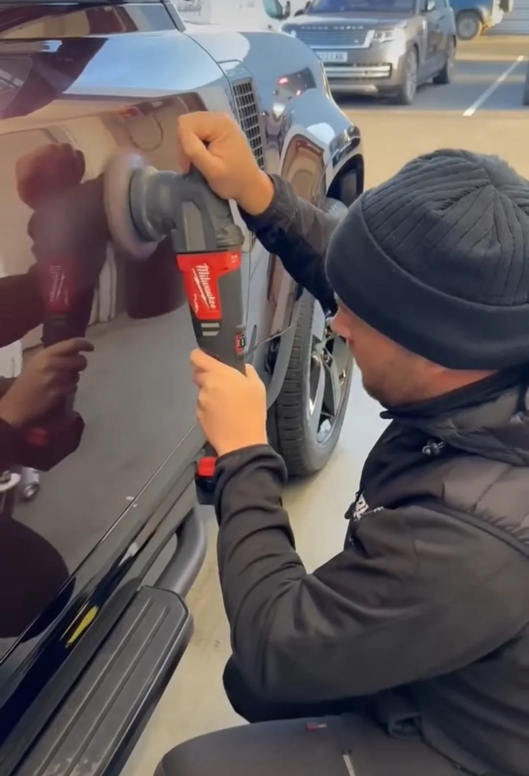 A person wearing a black beanie and jacket is polishing the side of a dark-colored car using a red electronic polisher, conveying focus and diligence.