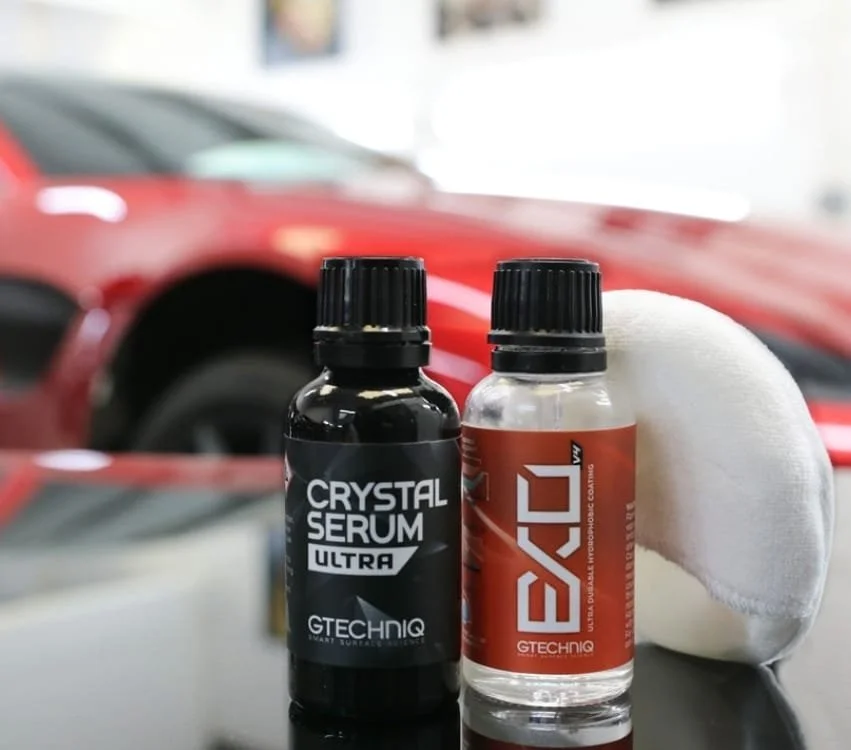 Two bottles labeled "Crystal Serum Ultra" and "EXO" stand on a reflective surface, with a red sports car and a white applicator pad in the background.