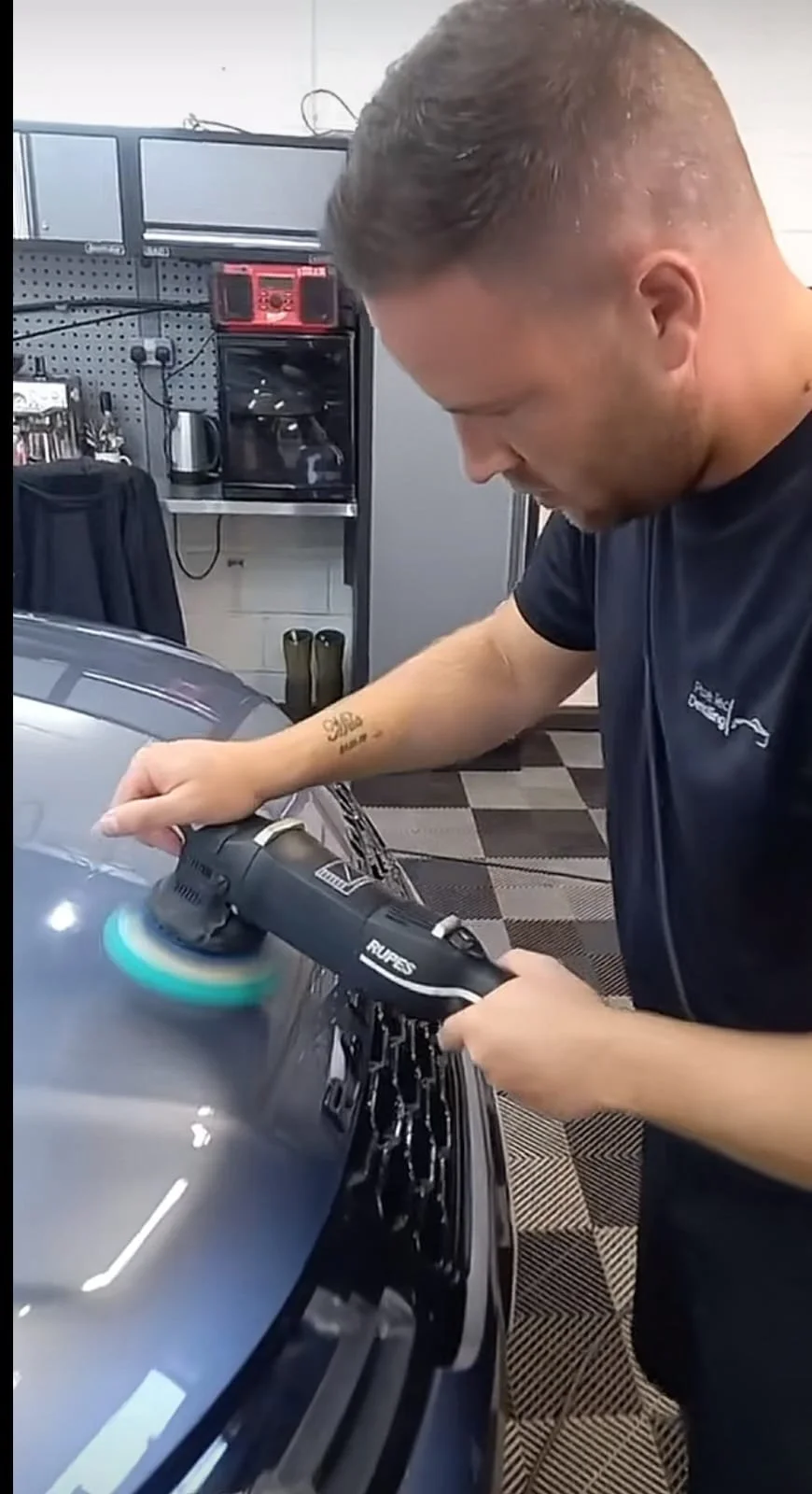 A man is using a car buffer on a vehicle's hood in a garage. He is focused on the task, with shelves and tools visible in the background.
