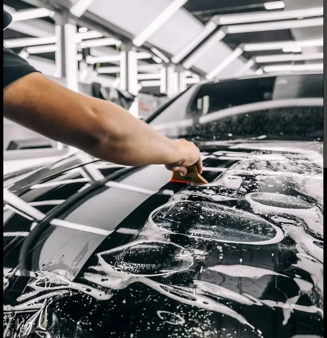 A person applies soapy water to a car hood in a well-lit garage, using a squeegee.