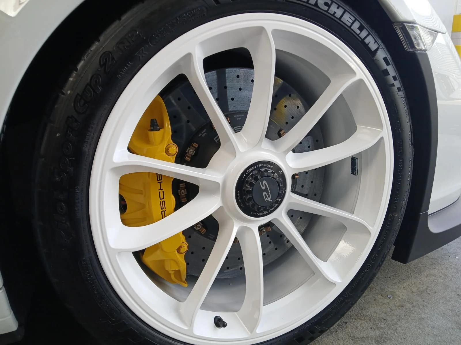 Close-up of a white car wheel with a prominently visible yellow brake caliper, sleek white spokes, a detailed central hub
