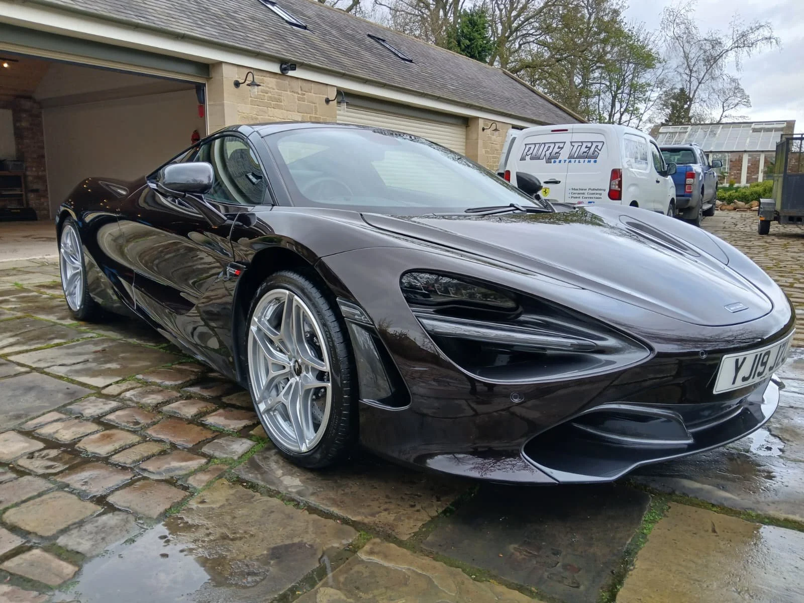 A sleek black sports car with glossy finish is parked on a wet stone driveway, in front of an open garage