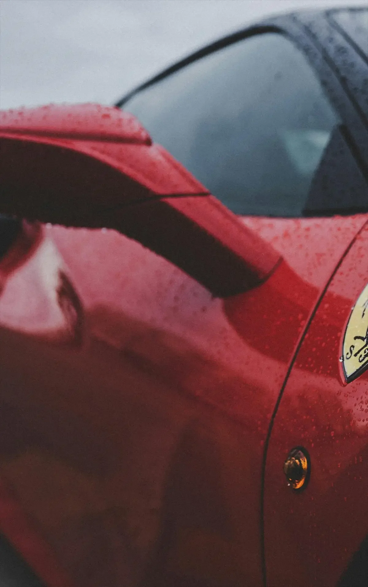 Close-up of a sleek red sports car with raindrops on the surface, showcasing a side mirror and emblem. The atmosphere is dynamic and luxurious.