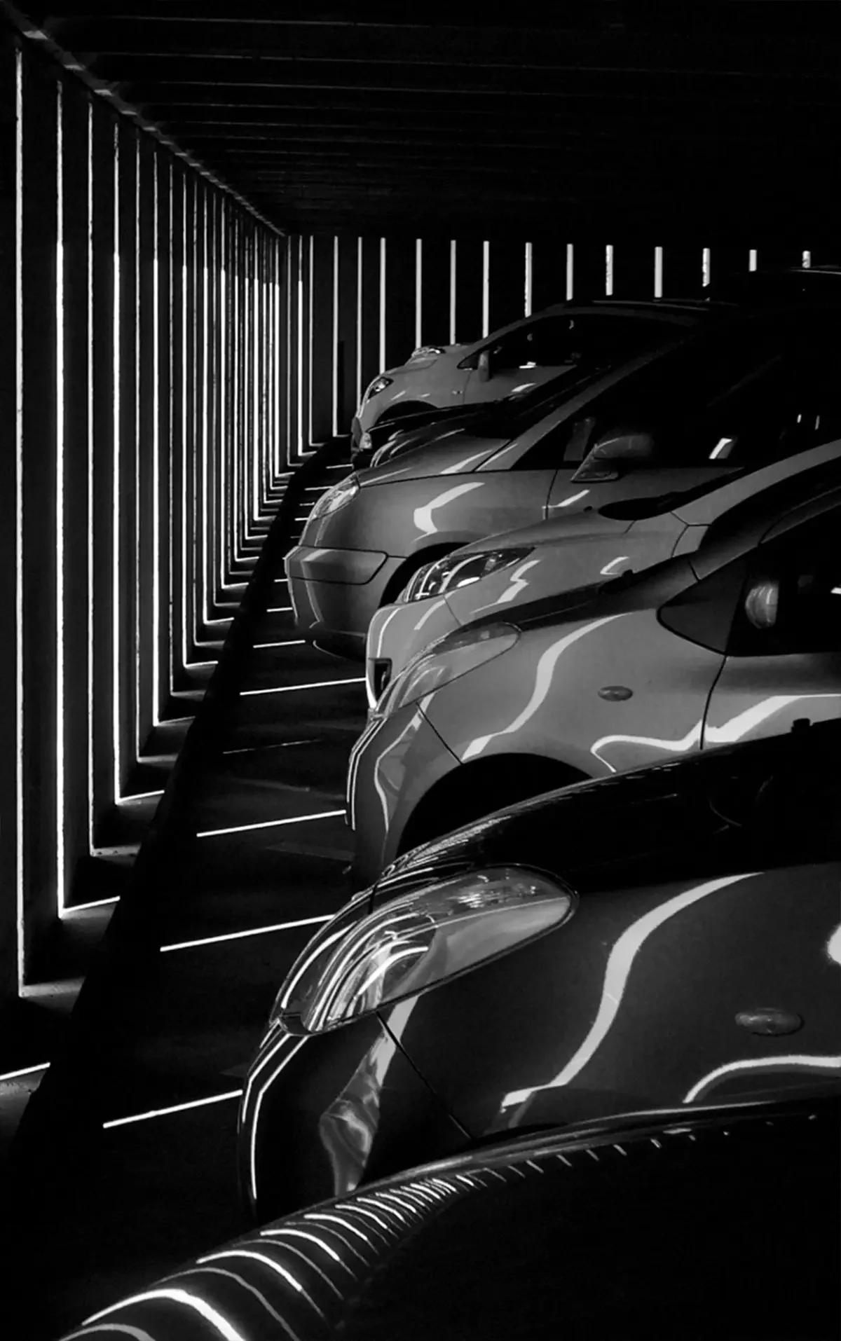 Black and white image of parked cars in a garage. Sunlight creates striking diagonal patterns of light and shadow, conveying a dramatic, abstract feel.
