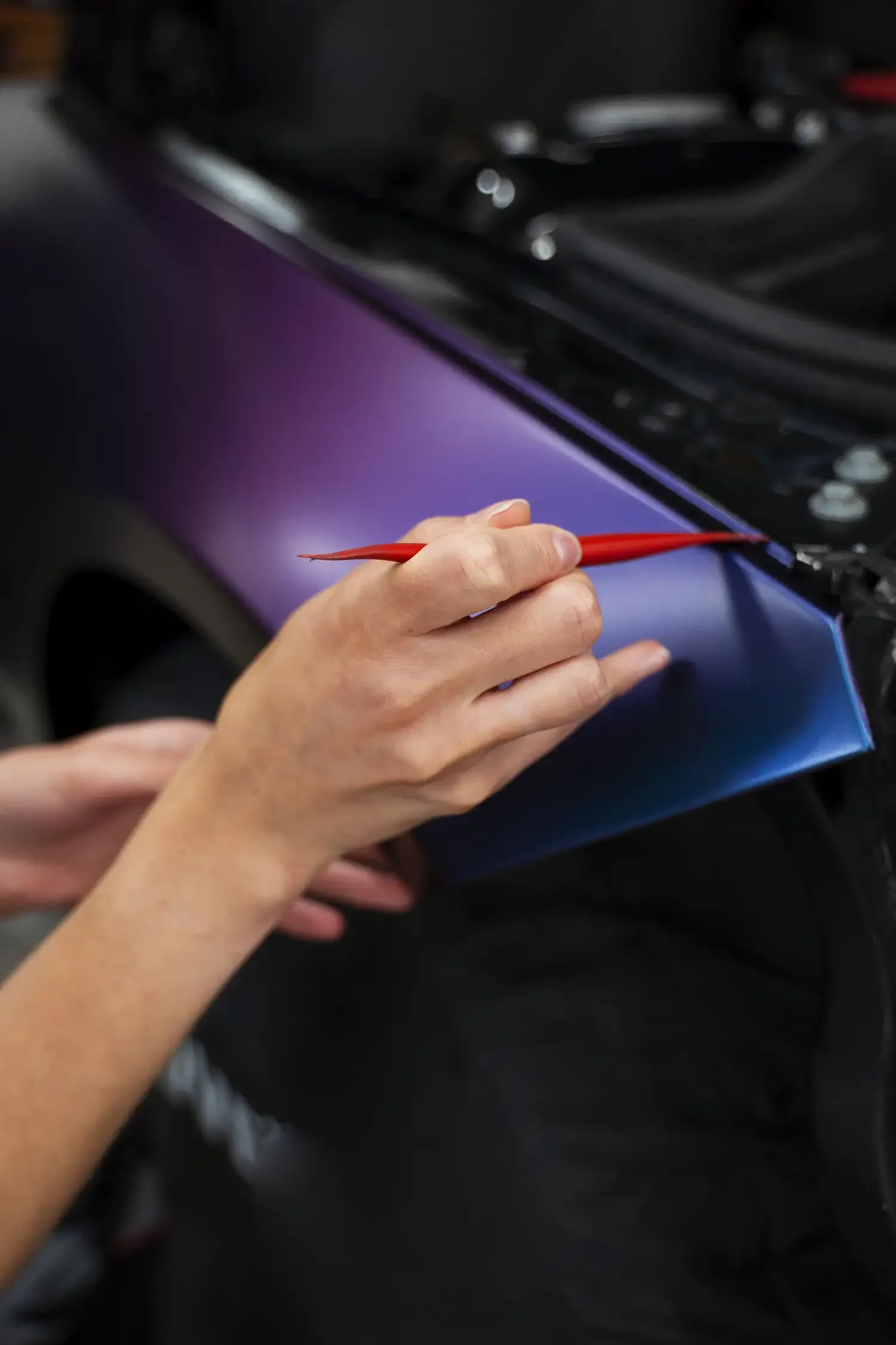 Hands apply a color-shifting vinyl wrap with a tool on a car fender, transitioning from purple to blue