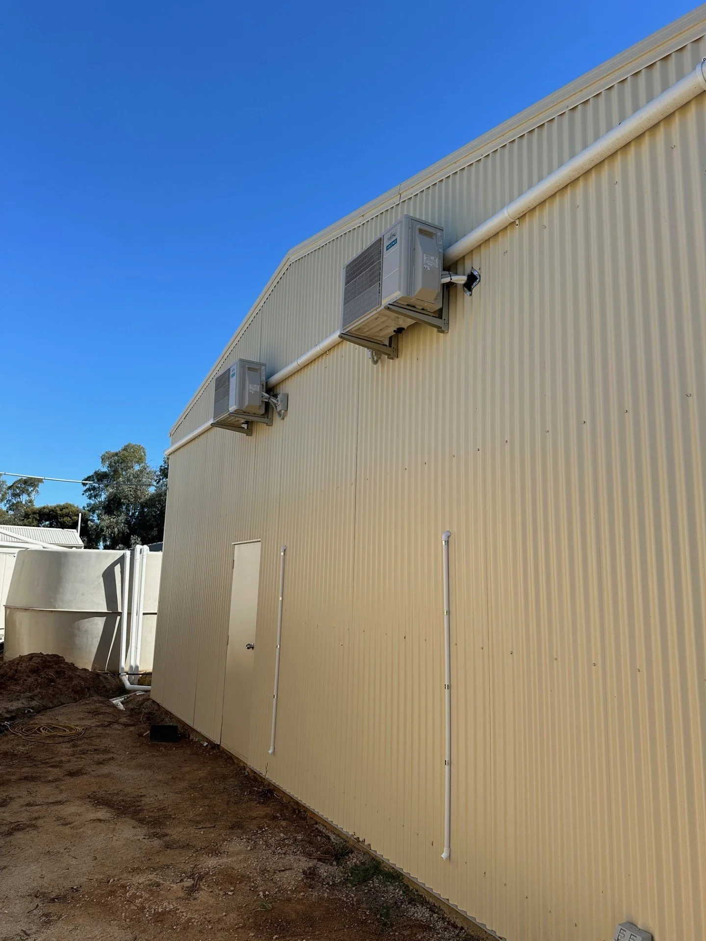 Side of a yellow industrial building with two wall-mounted air conditioning units and white conduit pipes running vertically along the wall.