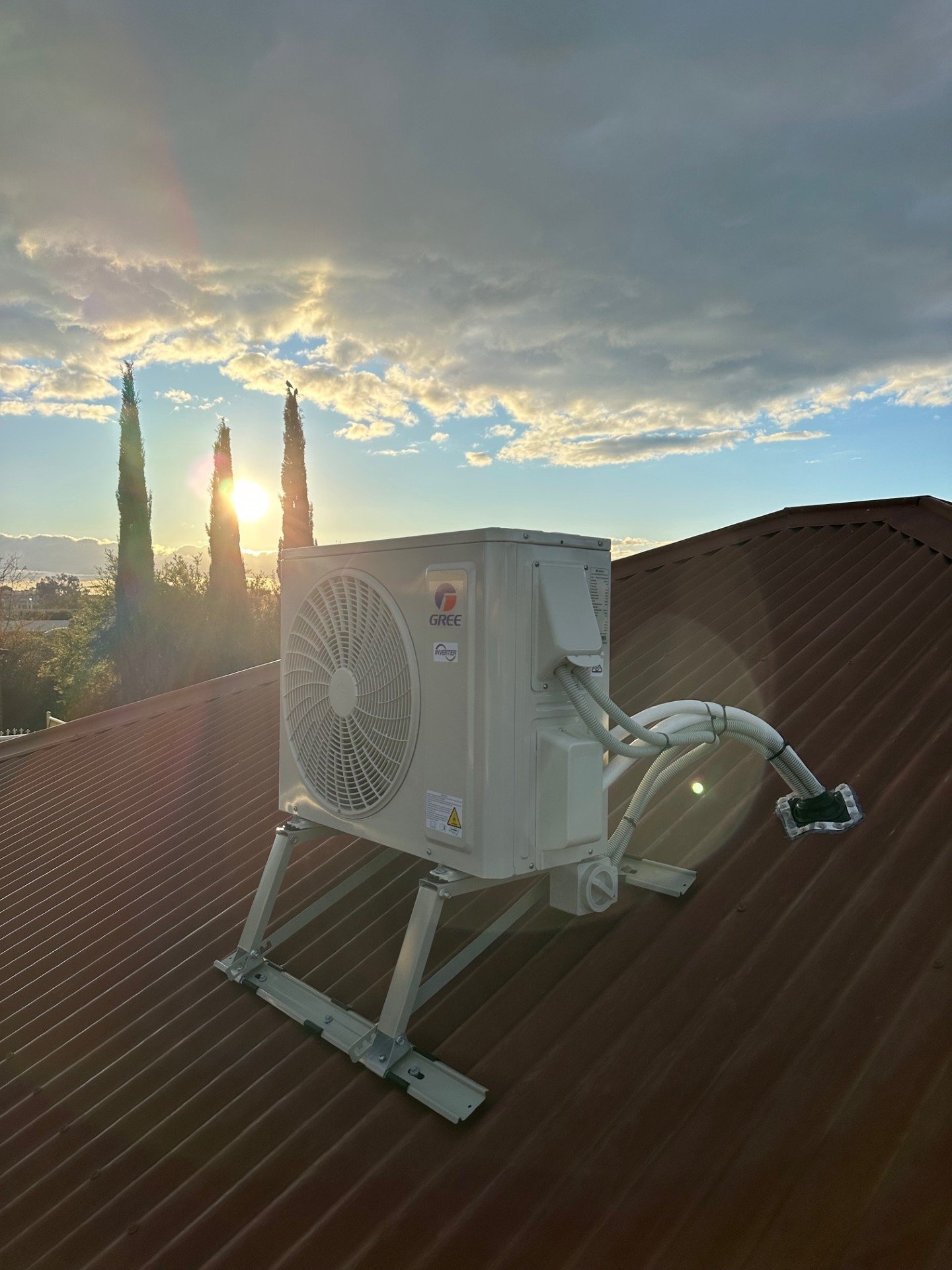 An air conditioning unit installed on a brown metal roof with trees and a setting sun in the background.