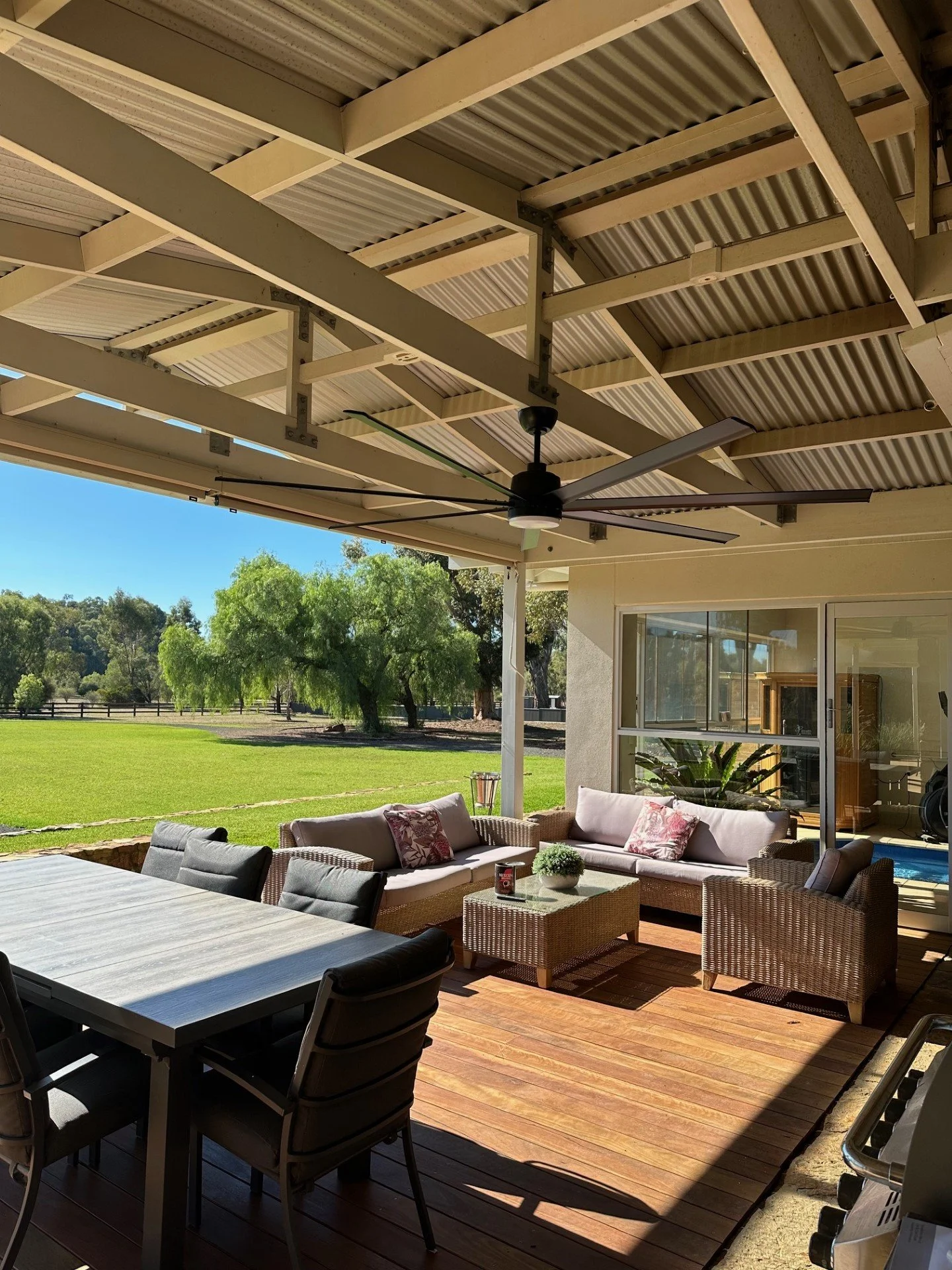Covered patio with outdoor furniture including a table, chairs, reach sofas, and potted plants, overlooking a grassy yard with trees.