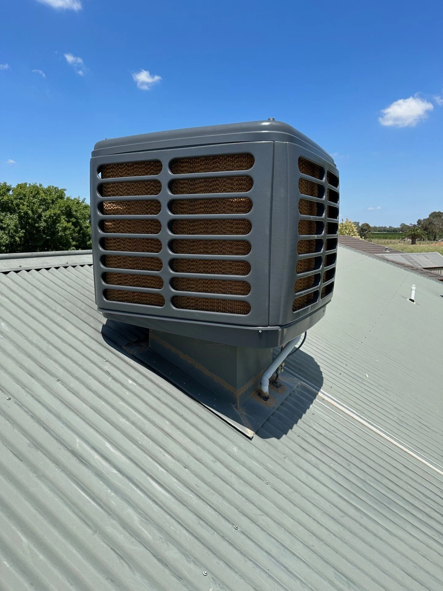 A rooftop air conditioning unit mounted on a metal roof, with a background of blue sky and trees.