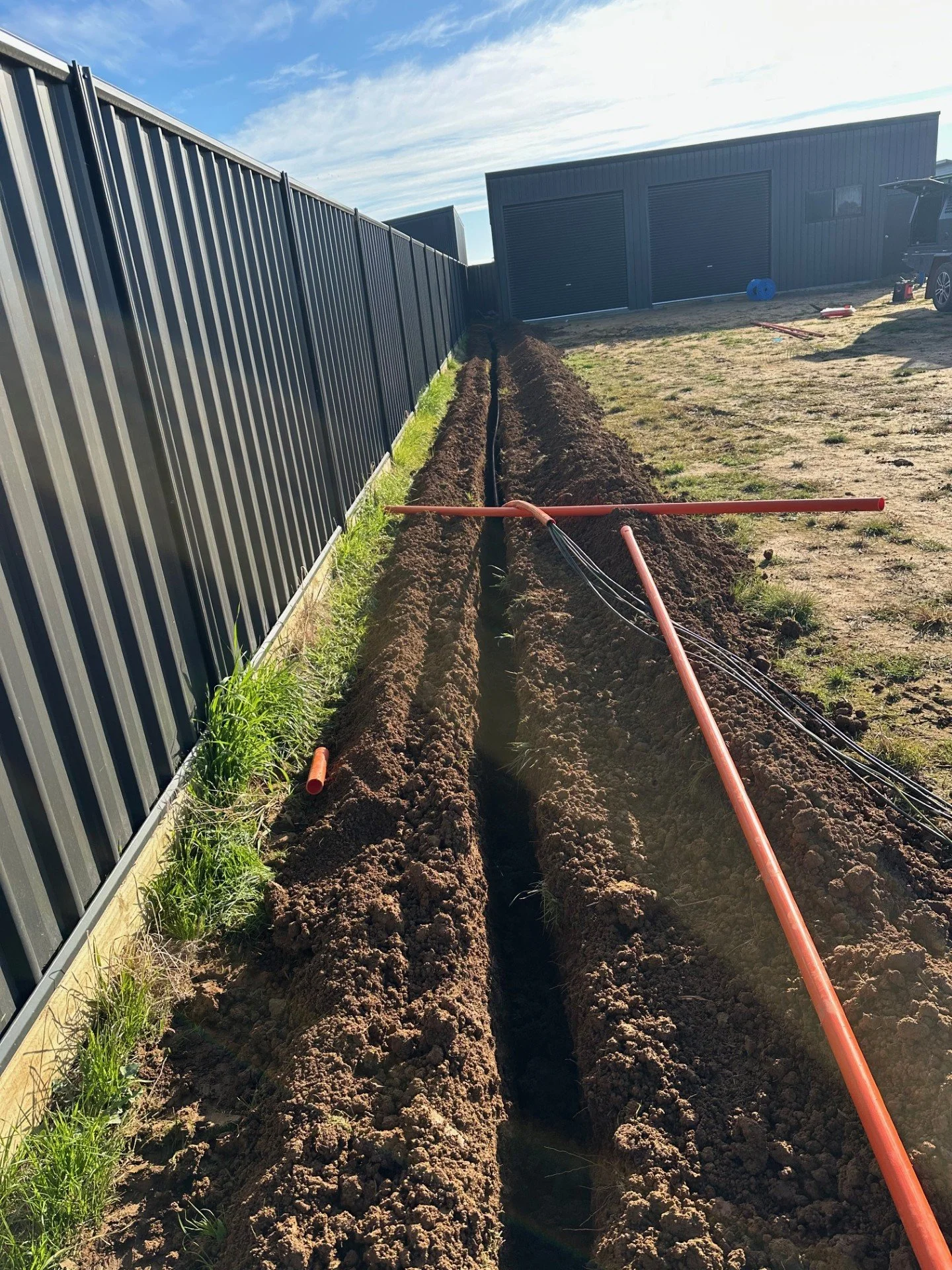 A trench with orange piping and black electrical cables being installed on a backyard with a black metal fence and a gray shed in the background.