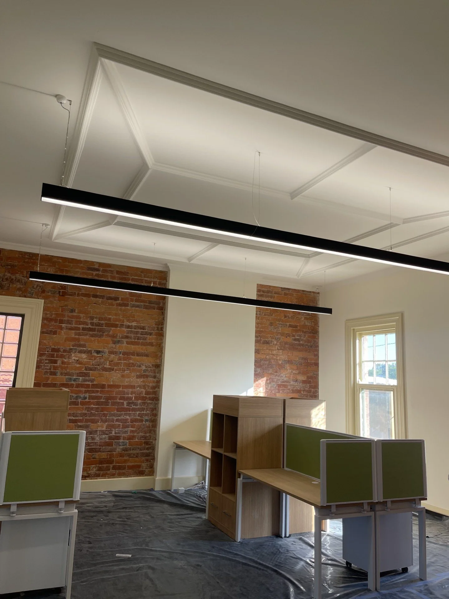 Modern office space with exposed brick walls, ceiling molding, large windows, and suspended linear light fixtures.